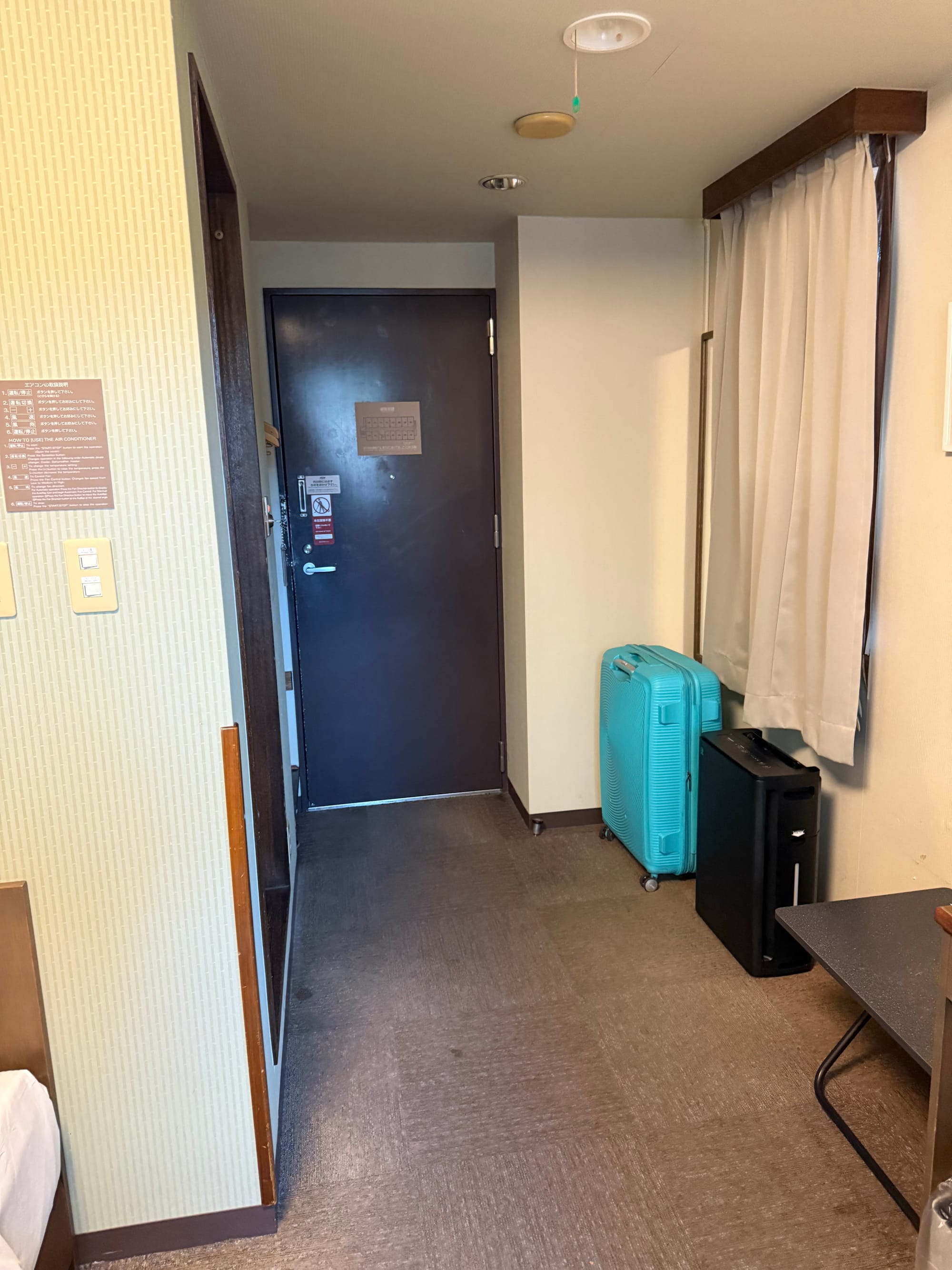 Guest room entrance area at Hotel Route-Inn Kamisuwa, showing the door, luggage, air purifier, small table and curtained window in a compact hotel room.