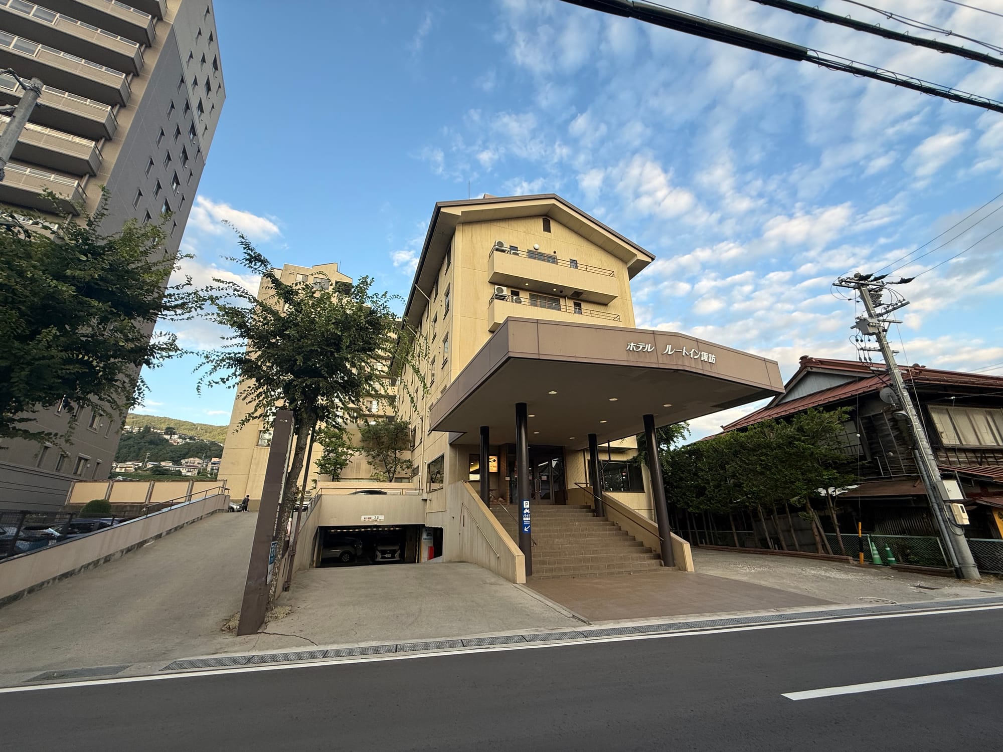 Hotel Route-Inn Kamisuwa building in Suwa, Nagano Prefecture, viewed from the street, with the main entrance canopy, surrounding buildings and a partly cloudy sky.