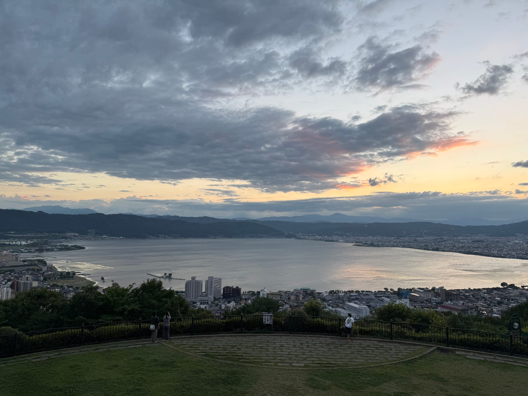 View of Lake Suwa from Tateishi Park at sunset, with the lakeshore town, surrounding mountains and dramatic clouds reflecting the evening light across the water.