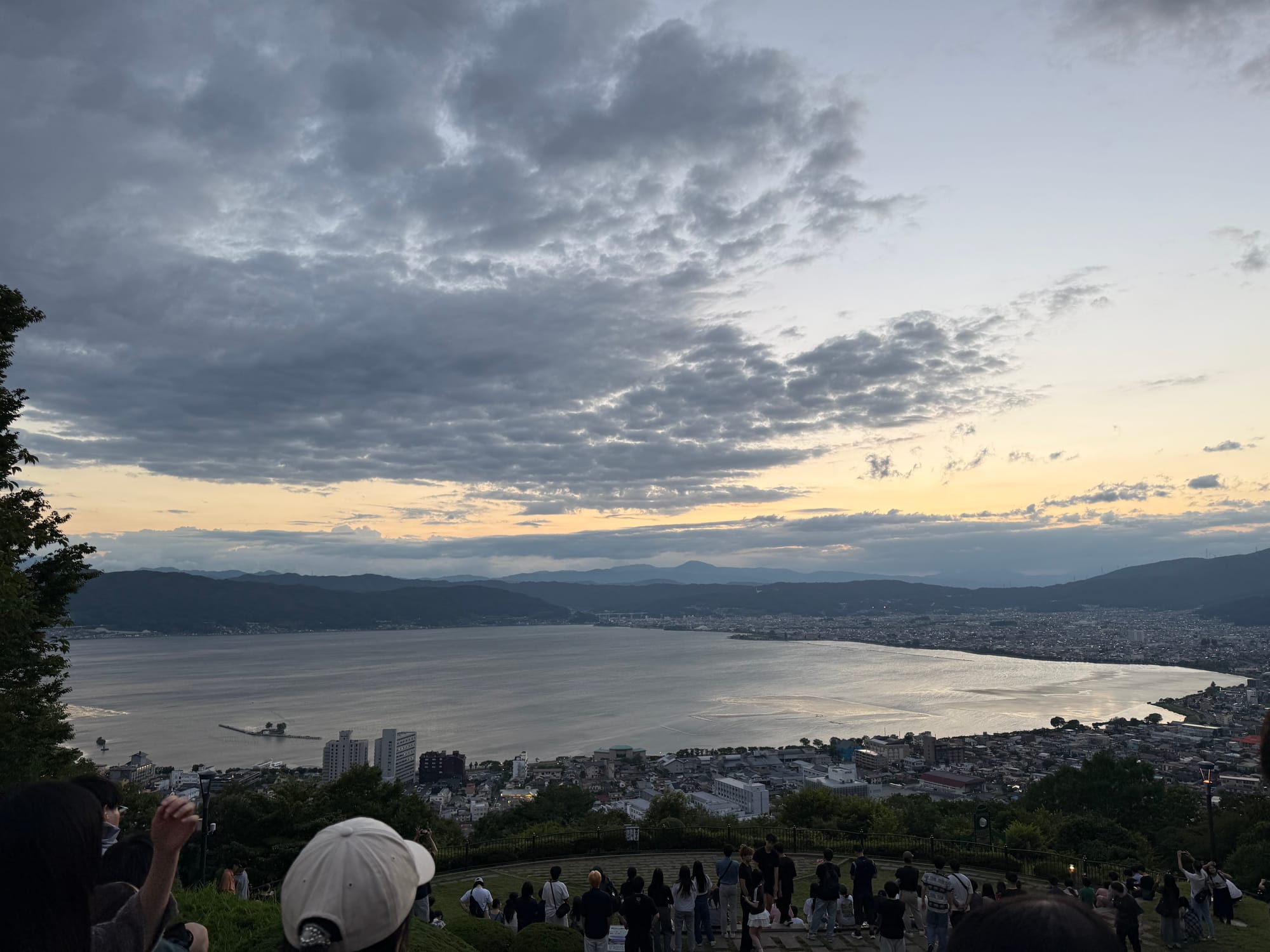 Visitors gather at Tateishi Park overlooking Lake Suwa at sunset, with the lake, surrounding towns and mountain ranges illuminated beneath a dramatic cloud-filled sky.