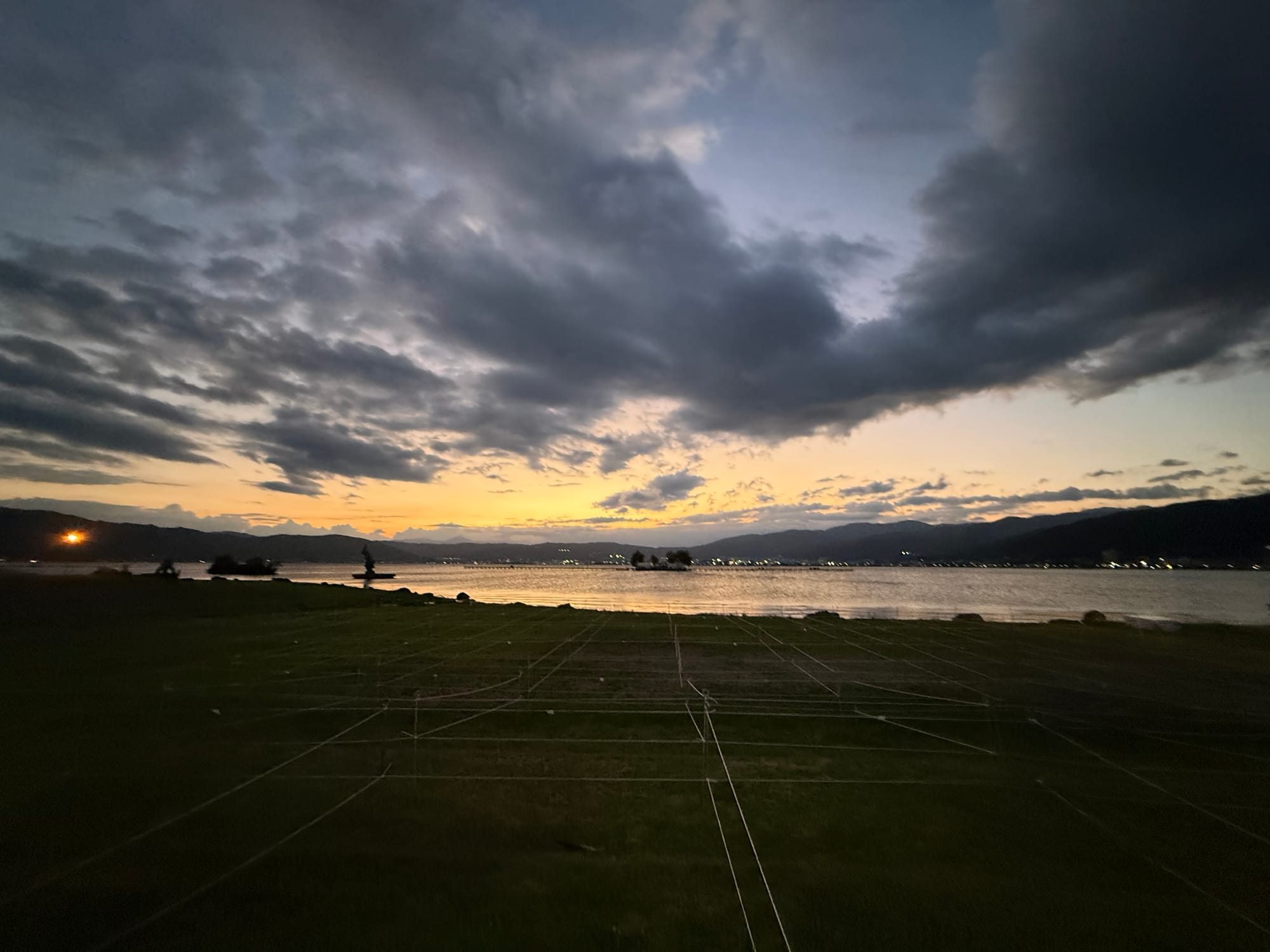 Sunset over Lake Suwa viewed from Suwa Lakeside Park, with dark clouds overhead, fading golden light on the water and mountains lining the distant shoreline.