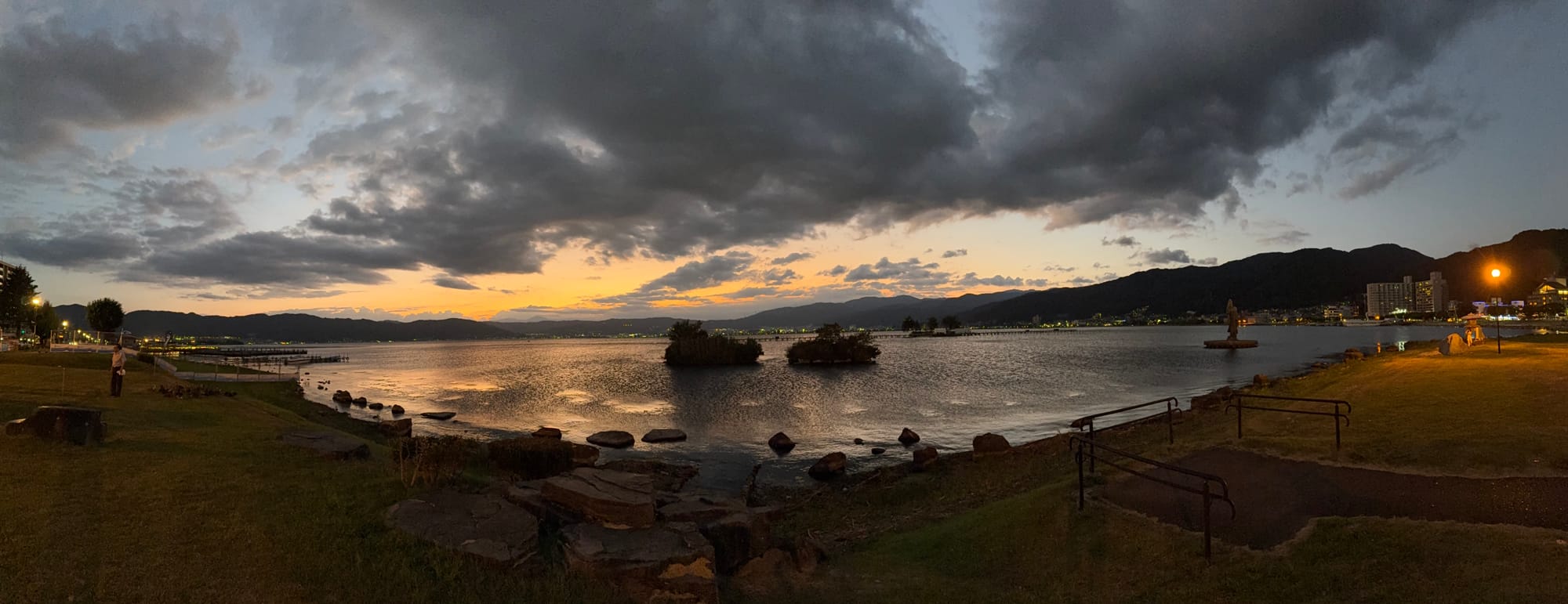 Panoramic evening view of Lake Suwa from Suwa Lakeside Park, showing small vegetated islets, shoreline lights, mountains and dramatic clouds above the water at sunset.