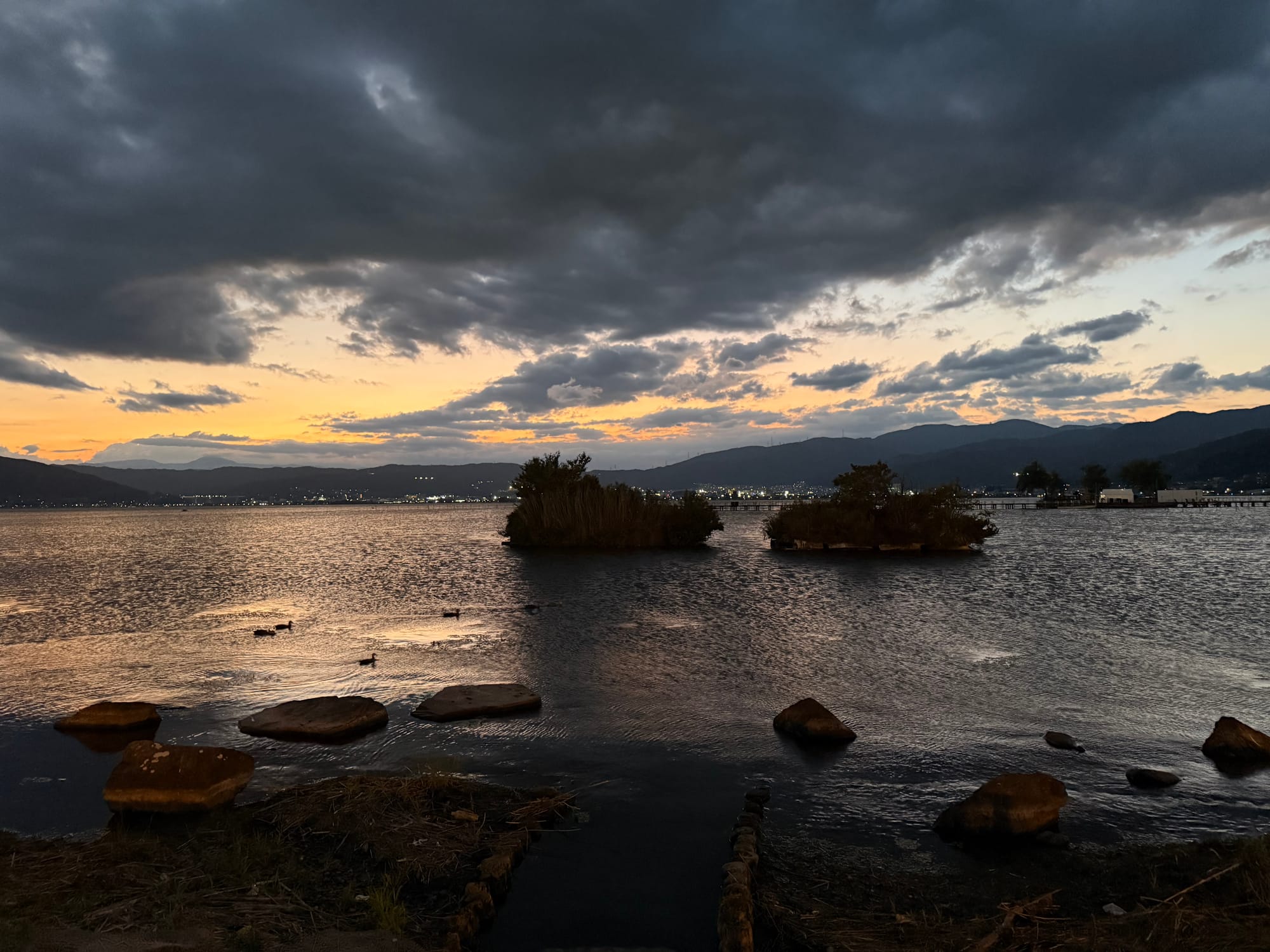 Twilight view across Lake Suwa, with small reed-covered islets, ducks on the water, rocky shoreline and dark storm clouds illuminated by the fading sunset.