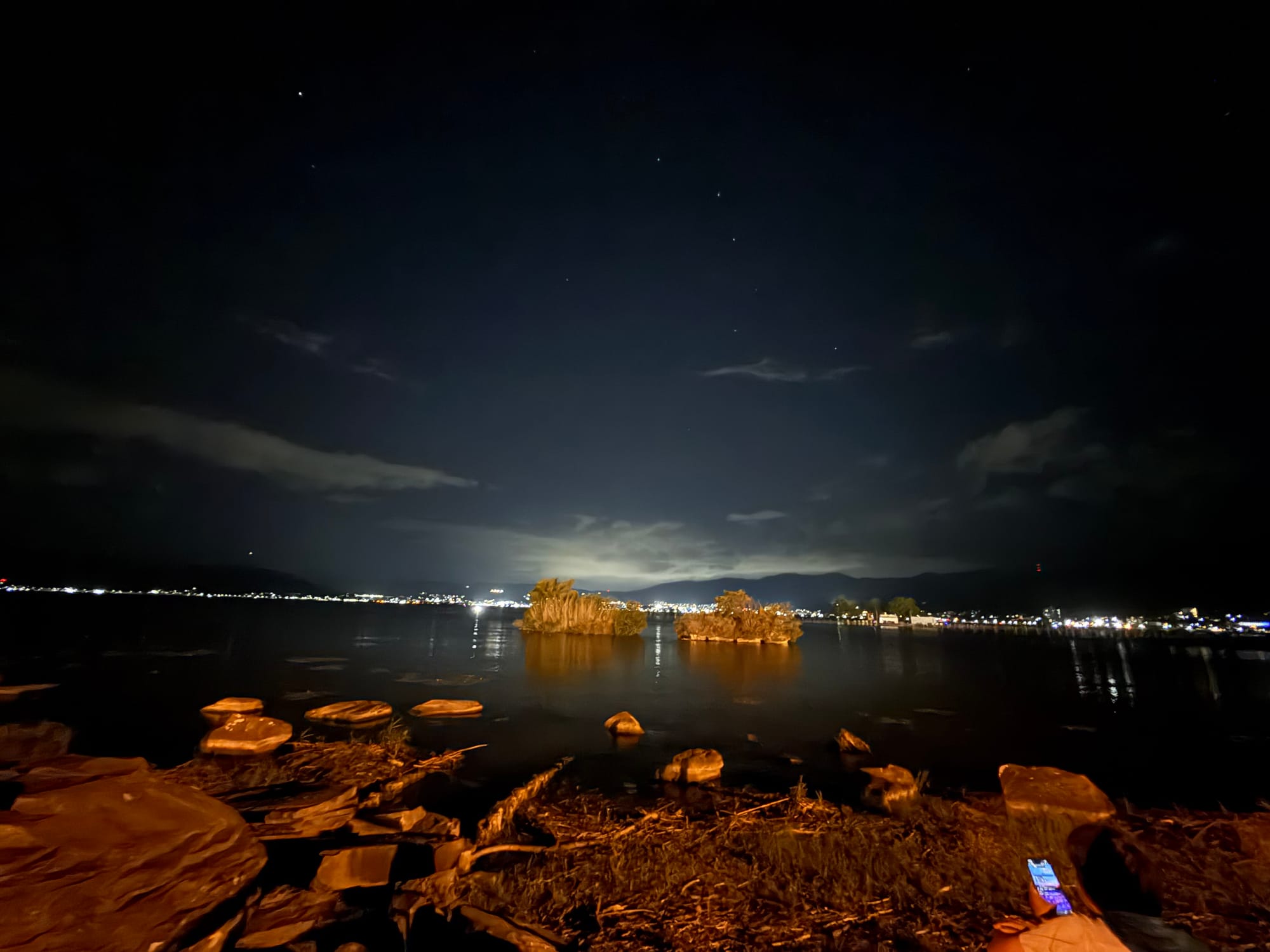 Night view of Lake Suwa before a fireworks display, with illuminated reed-covered islets, calm water, shoreline lights, rocky foreground and stars visible between clouds.