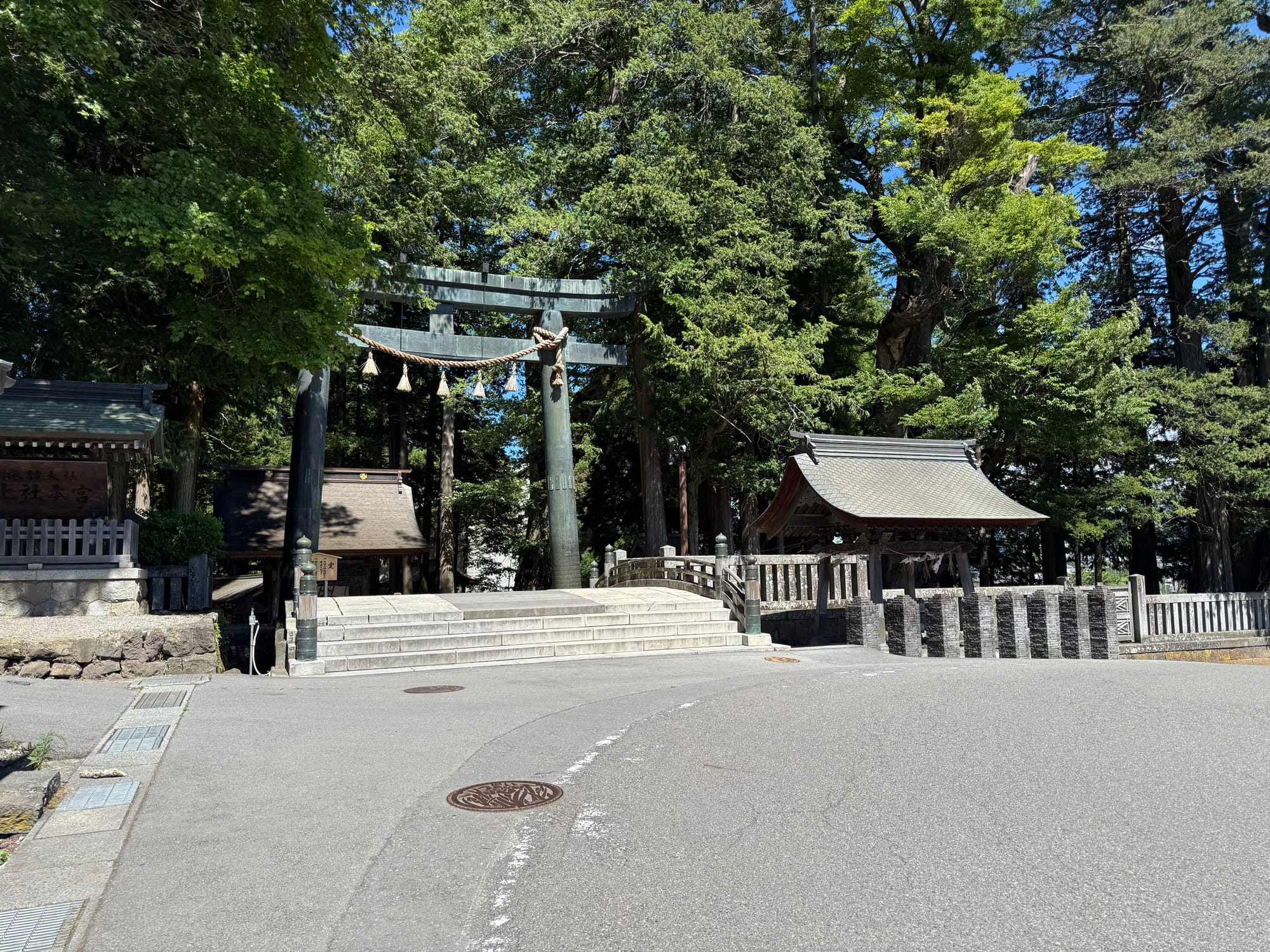 Main torii gate of Suwa Taisha Kamisha Honmiya, surrounded by towering cedar trees, with stone steps, shrine buildings and sacred rope decorations at the entrance.