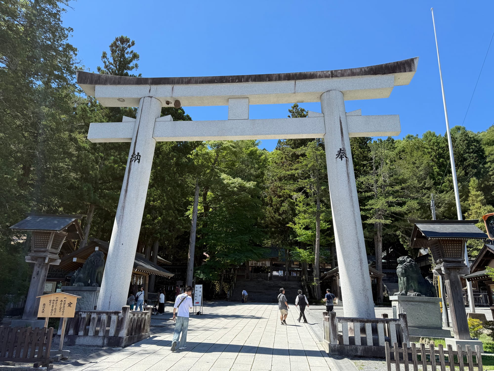 Large stone torii gate at Suwa Taisha Kamisha Honmiya, framing the shrine approach with visitors, guardian lion-dog statues and a forest of towering cedar trees beyond.