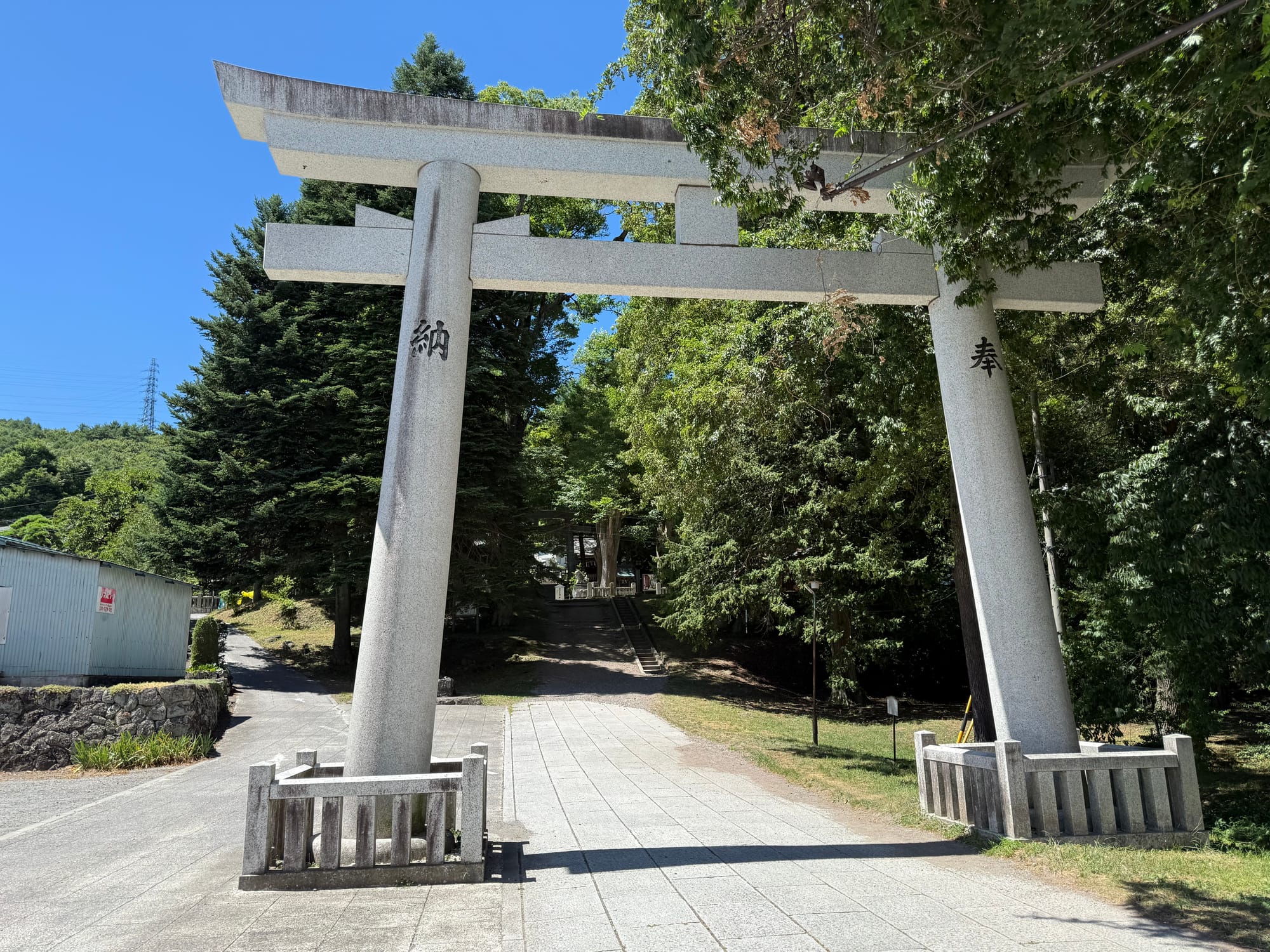 Stone torii gate at Suwa Taisha Kamisha Maemiya, marking the approach to the shrine through a wooded setting, with a pathway leading toward the main shrine buildings.