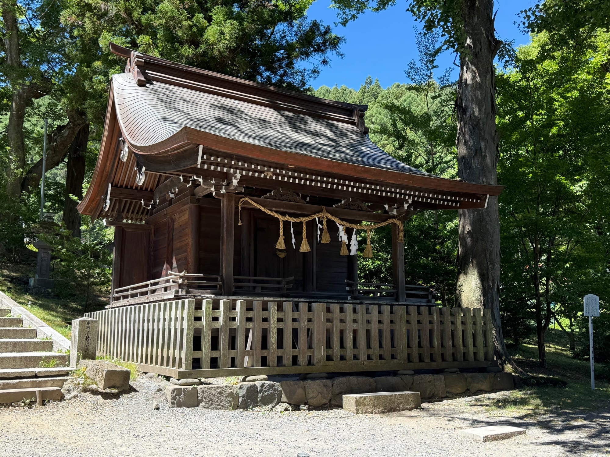 Traditional wooden shrine building at Suwa Taisha Kamisha Maemiya, surrounded by tall trees and enclosed by a wooden fence within the shrine's forested grounds.