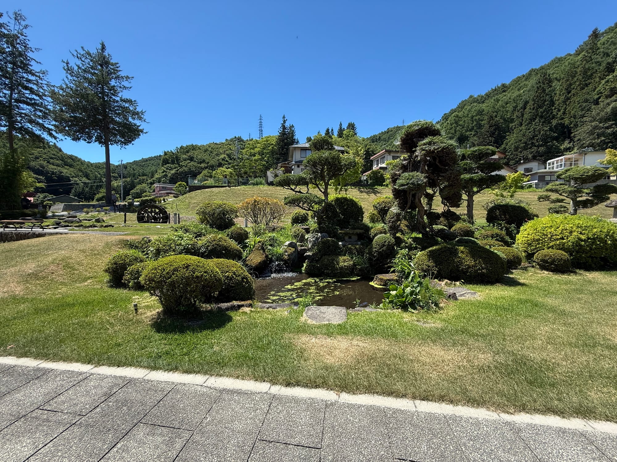 Landscaped Japanese garden with a small pond, carefully pruned shrubs and rolling lawns, set against forested hills and mountains under a clear blue sky.
