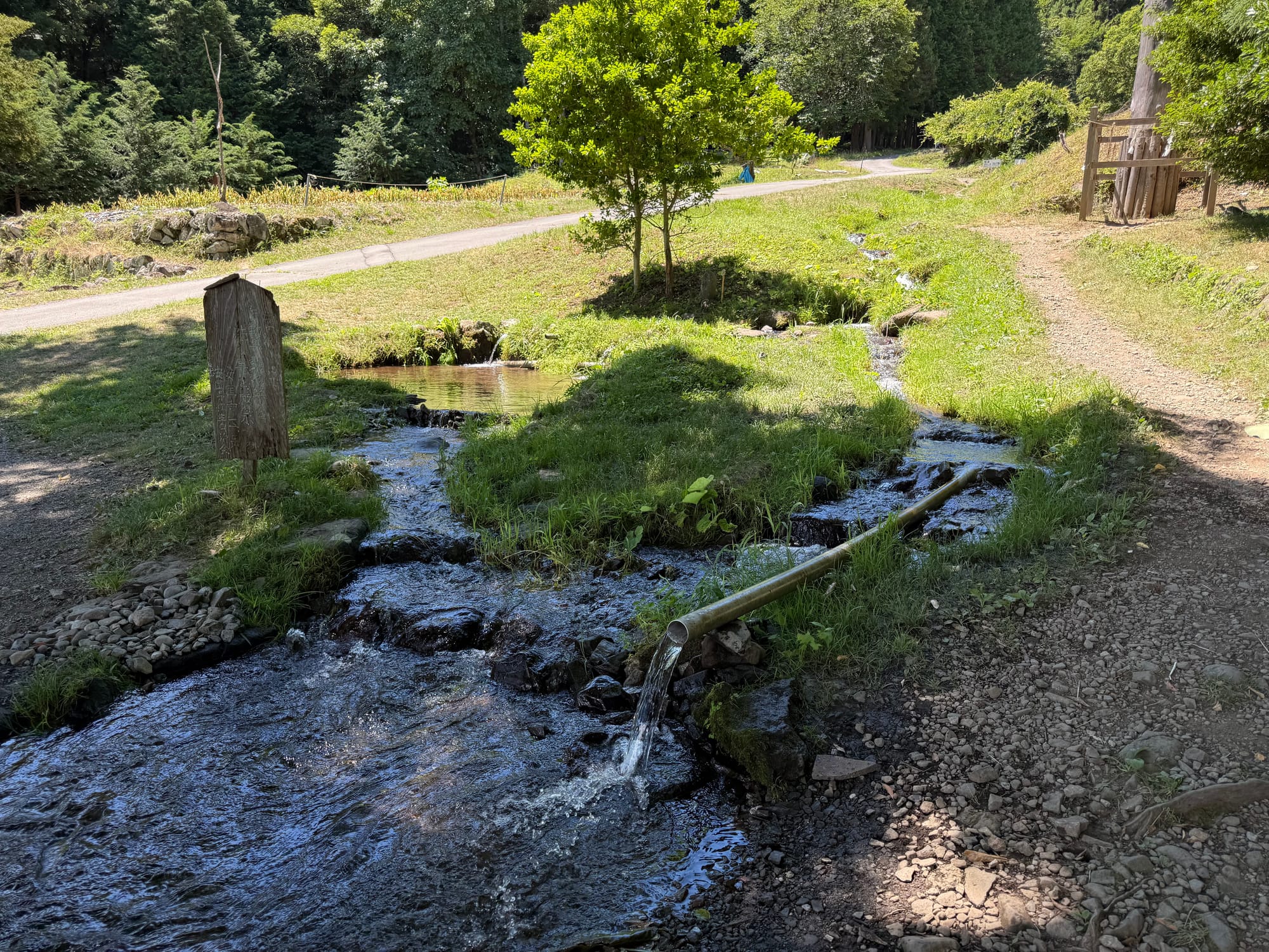 Small stream flowing through the grounds of Suwa Taisha Kamisha Maemiya, with clear water, grassy banks, a bamboo water channel and wooded hills in the background.