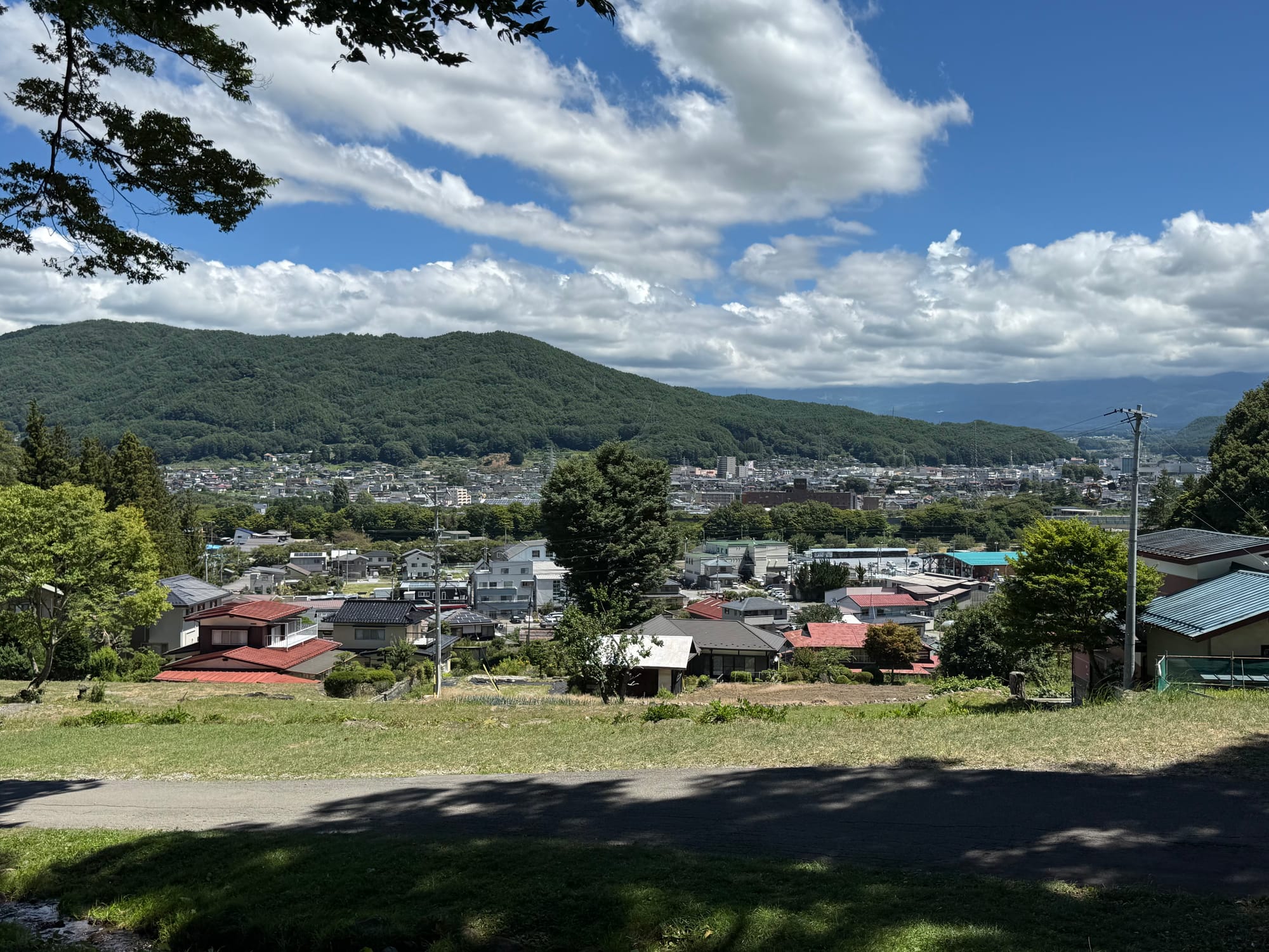 View from Suwa Taisha Kamisha Maemiya overlooking a valley town, with houses, forested hills and distant mountains beneath a sky filled with large summer clouds.