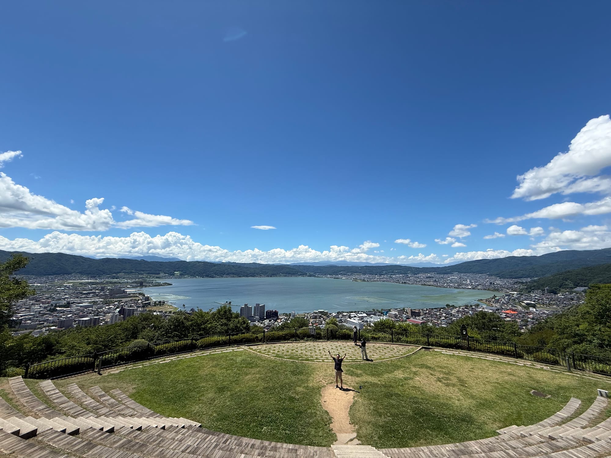 Panoramic view from Tateishi Park overlooking Lake Suwa, with the lakeside towns, surrounding mountains and an open observation terrace beneath a clear summer sky.