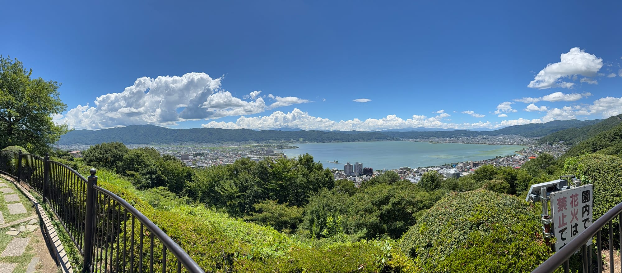 Panoramic view of Lake Suwa from Tateishi Park, overlooking the lake, surrounding towns and mountains beneath a clear blue sky with large summer clouds.