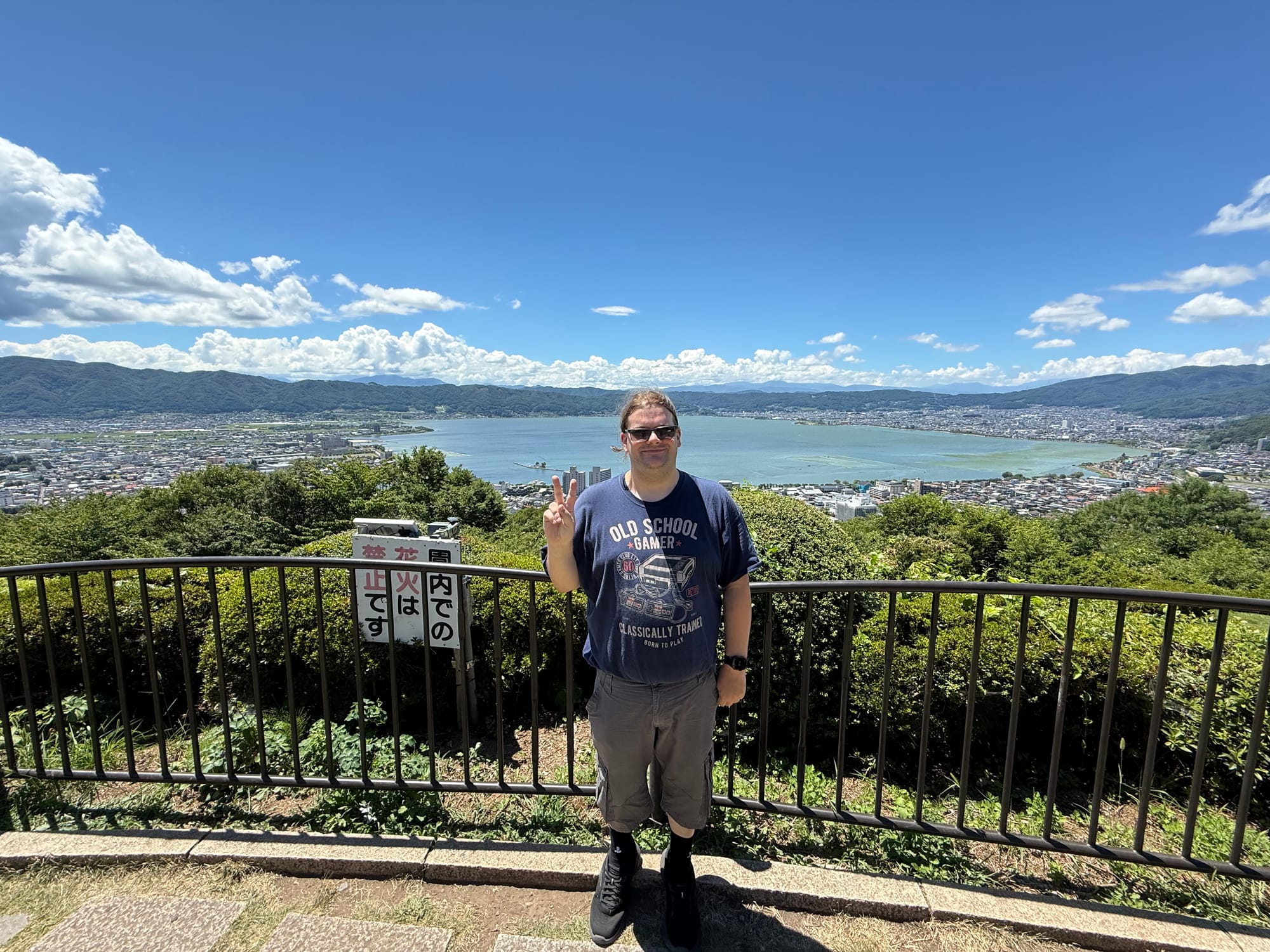 Visitor standing at the Tateishi Park viewpoint overlooking Lake Suwa, with the lake, surrounding towns and mountains visible beneath a clear blue summer sky.