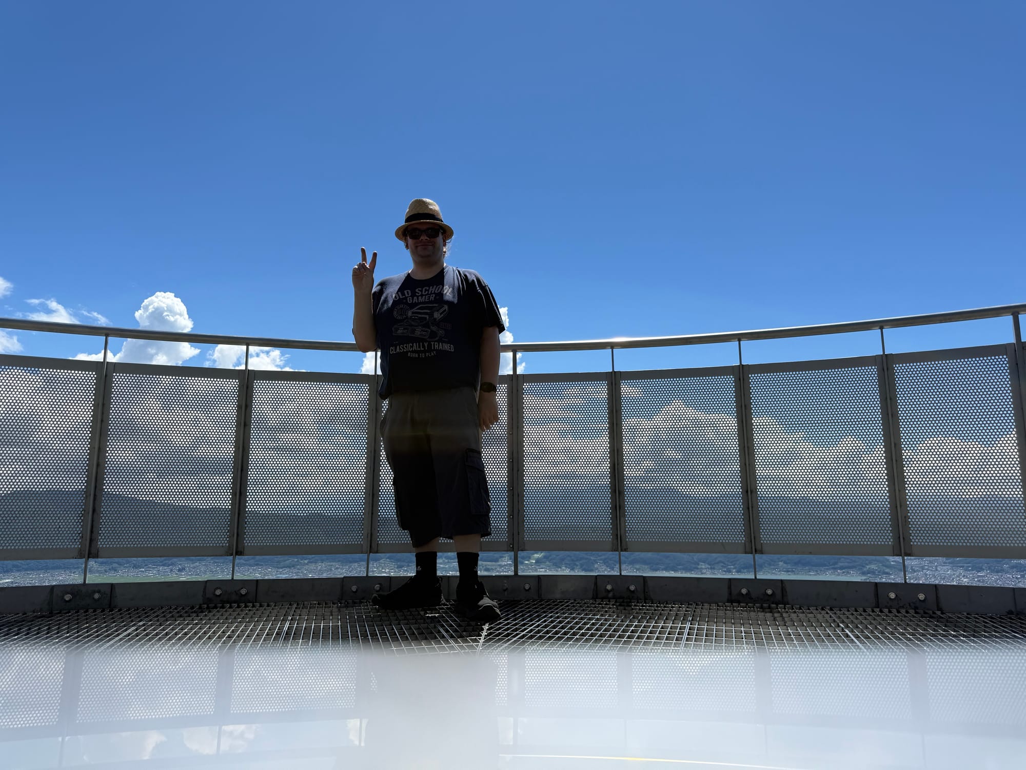 Visitor standing on the viewing platform at Oniyama Observation Point, with Lake Suwa and the surrounding mountains visible beyond the metal railing under a clear blue sky.