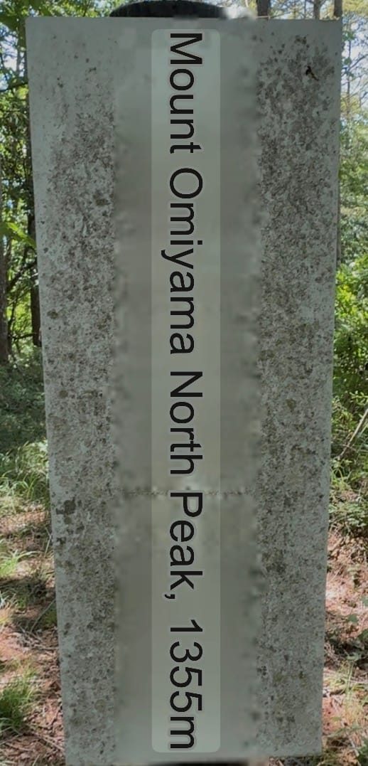 Stone summit marker at Mount Oniyama North Peak, indicating an elevation of 1,355 metres, standing beside a forest trail within the wooded mountains above Lake Suwa.