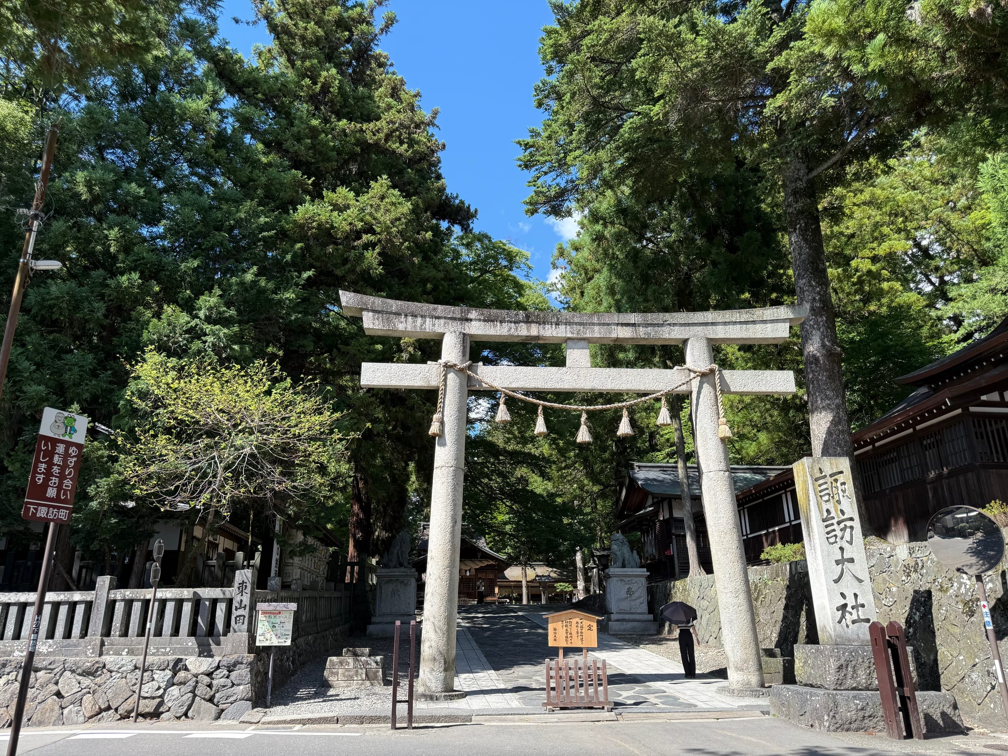 Stone torii gate at Suwa Taisha Shimosha Harumiya, marking the entrance to the shrine grounds, with towering cedar trees, shrine buildings and a paved approach beyond.
