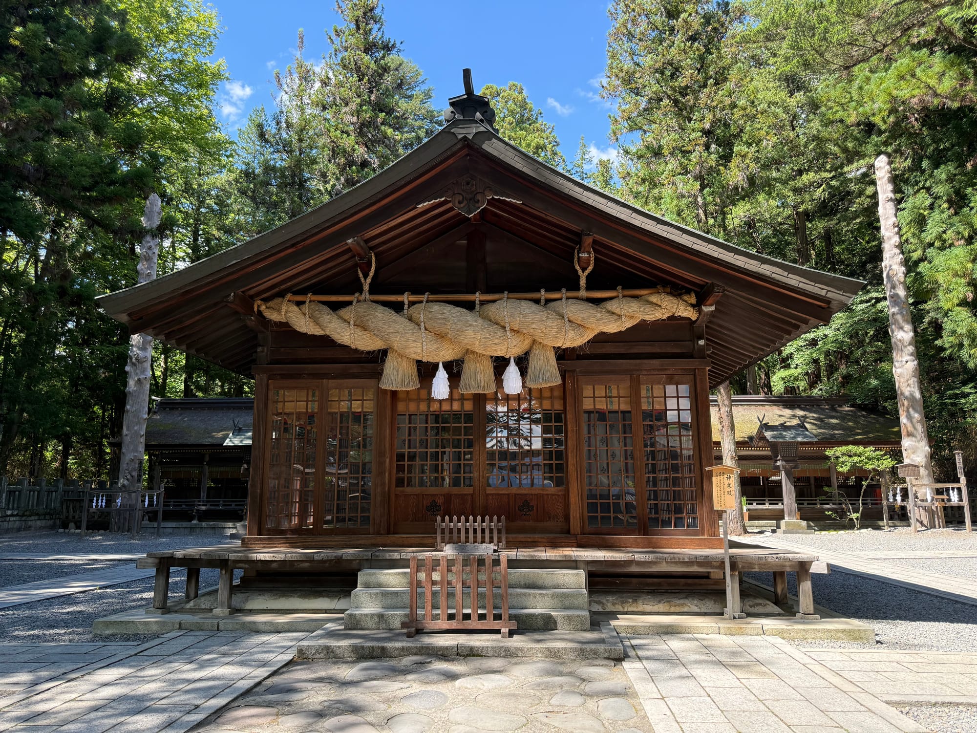 Wooden shrine building at Suwa Taisha Shimosha Harumiya, featuring a large sacred shimenawa rope, traditional architecture and towering cedar trees in the background.