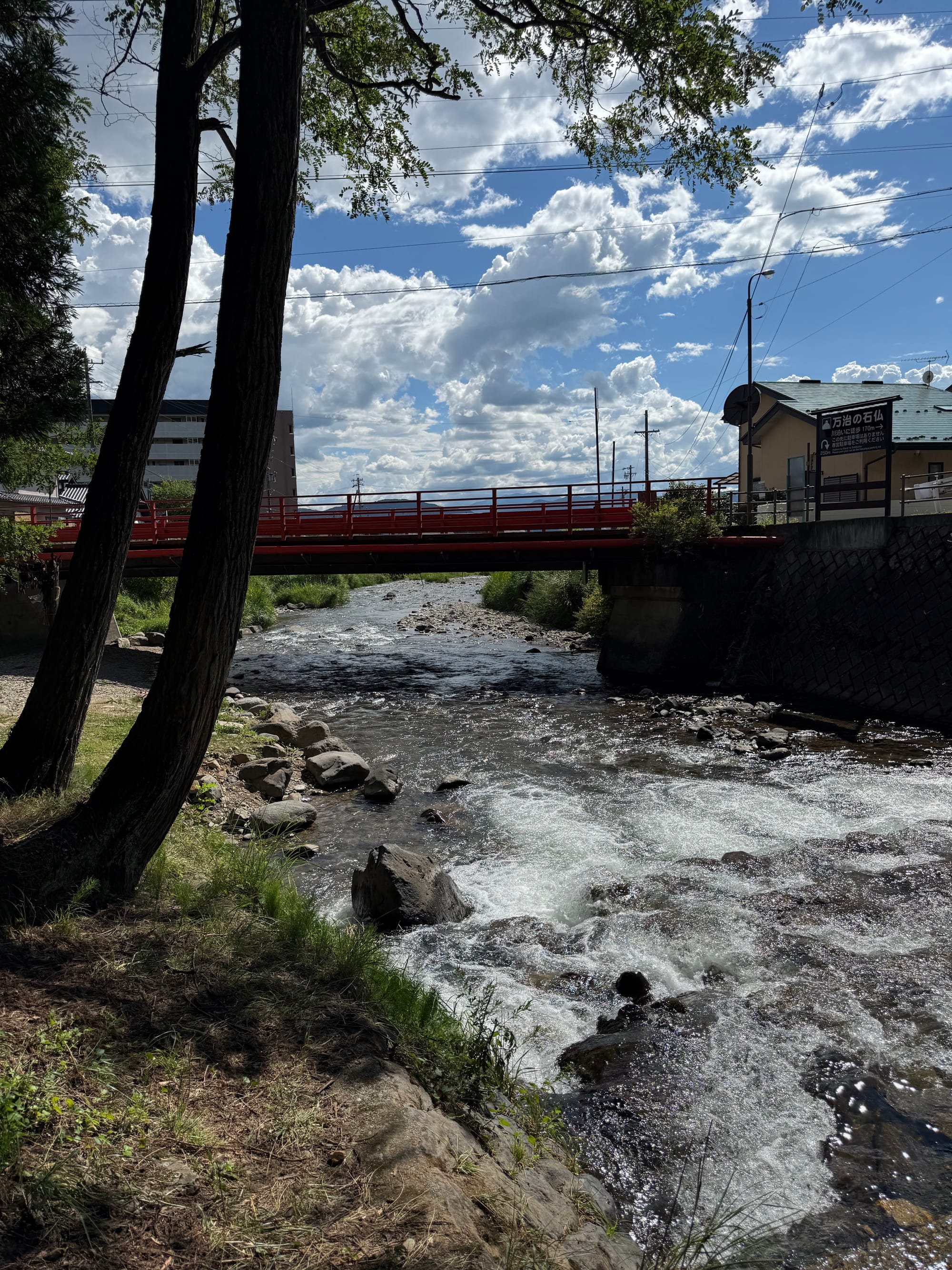 Mountain stream flowing beneath a red bridge near Suwa Taisha Shimosha Harumiya, with rocky banks, rushing water, nearby buildings and dramatic summer clouds overhead.