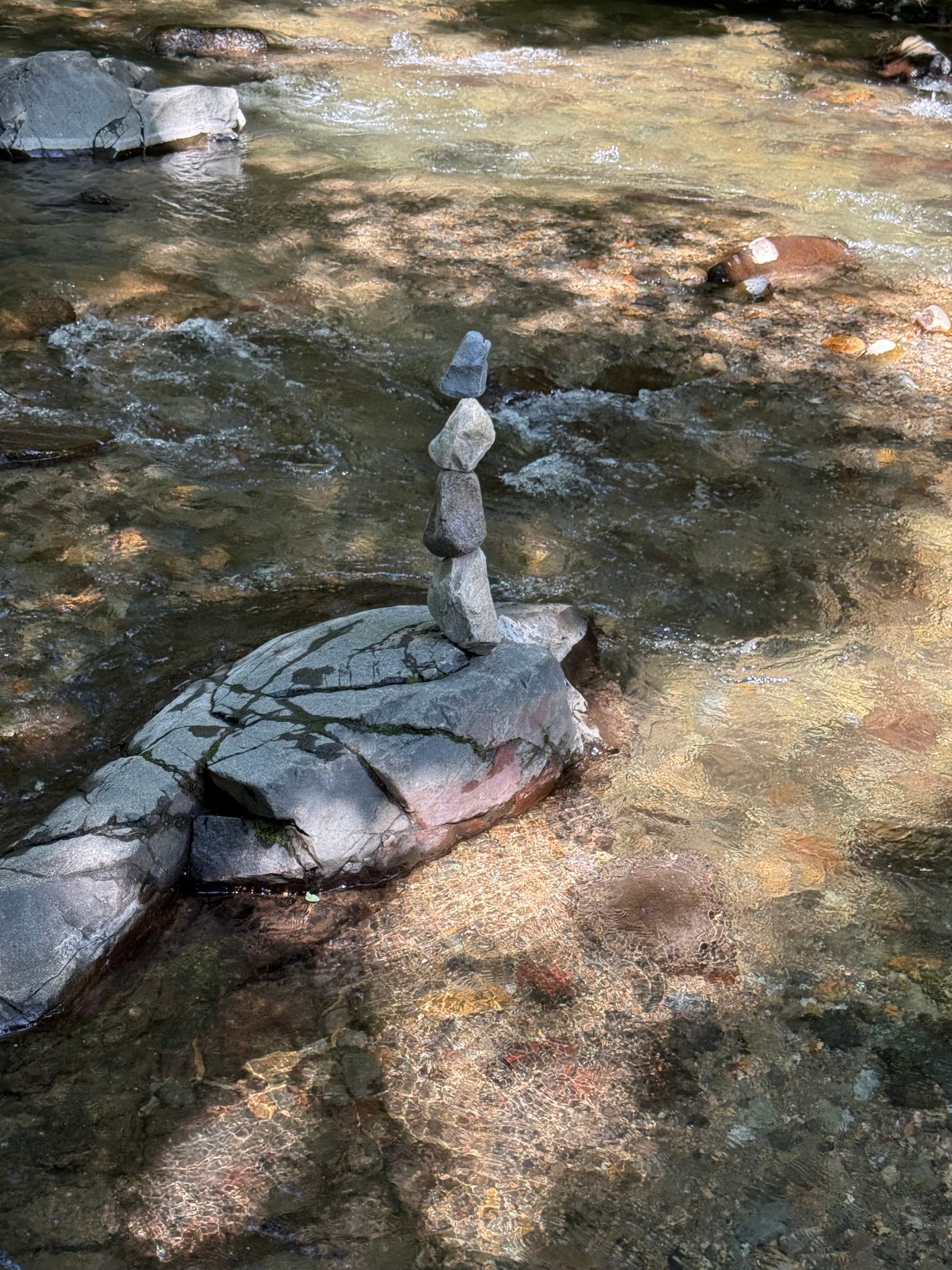 Stack of balanced stones on a rock in a clear mountain stream near Suwa Taisha Shimosha Harumiya, with sunlight reflecting through the shallow water and riverbed below.