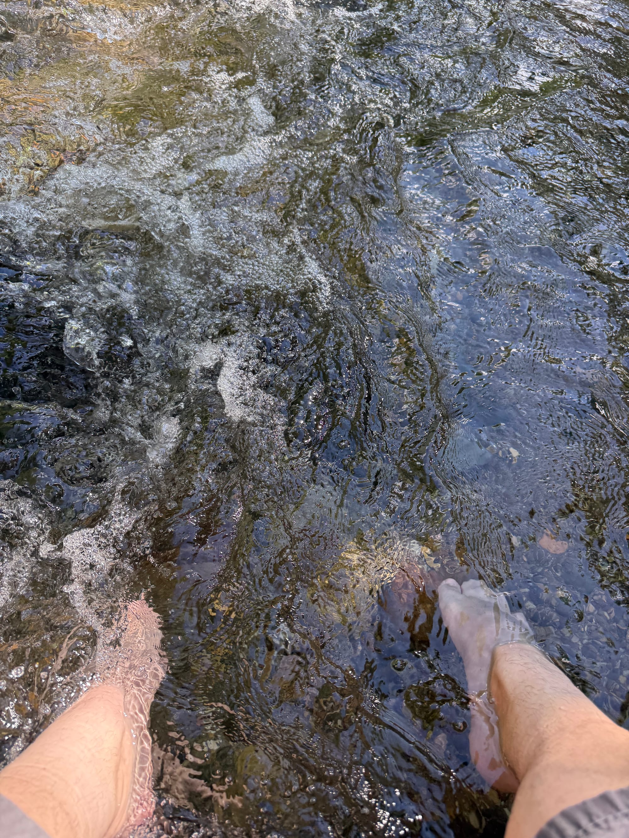 Bare feet submerged in a clear mountain stream near Suwa Taisha Shimosha Harumiya, with flowing water and rounded stones visible beneath the surface.
