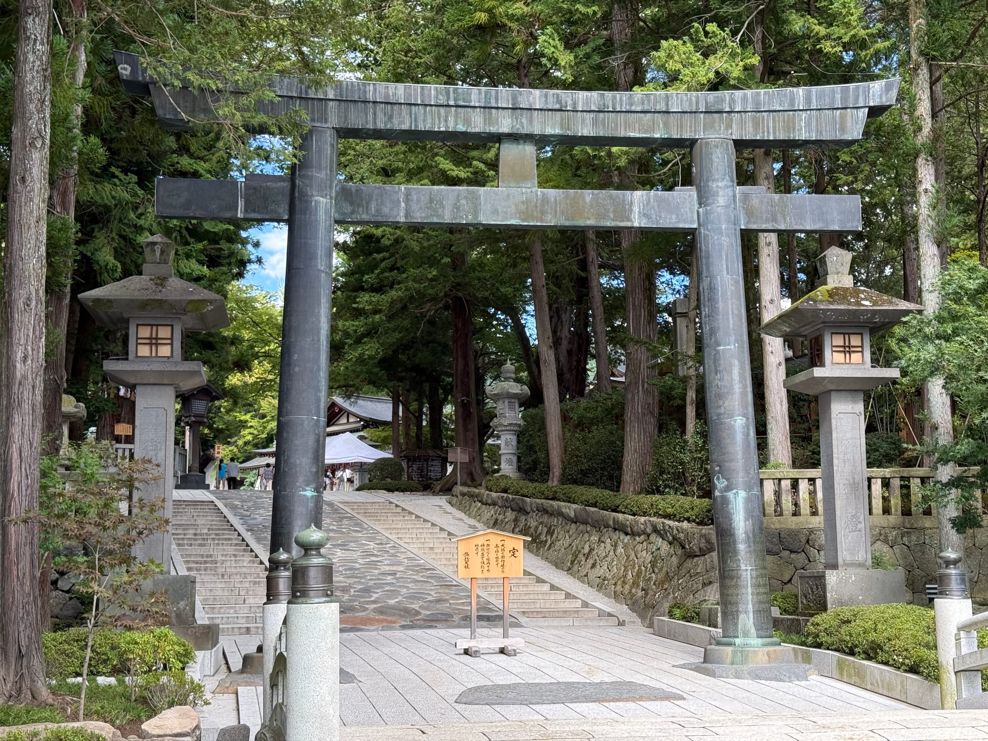 Bronze torii gate at Suwa Taisha Shimosha Akimiya, standing at the base of a stone approach lined with lanterns and towering cedar trees leading into the shrine grounds.