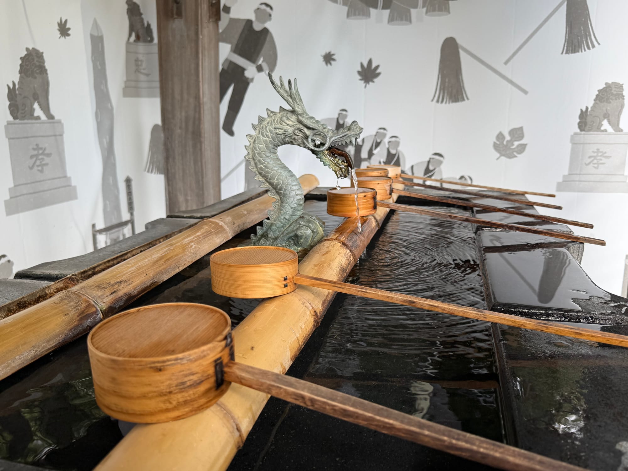 Dragon-shaped purification fountain at Suwa Taisha Shimosha Akimiya, with water flowing into a stone basin lined with bamboo rails and wooden ladles for ritual cleansing.
