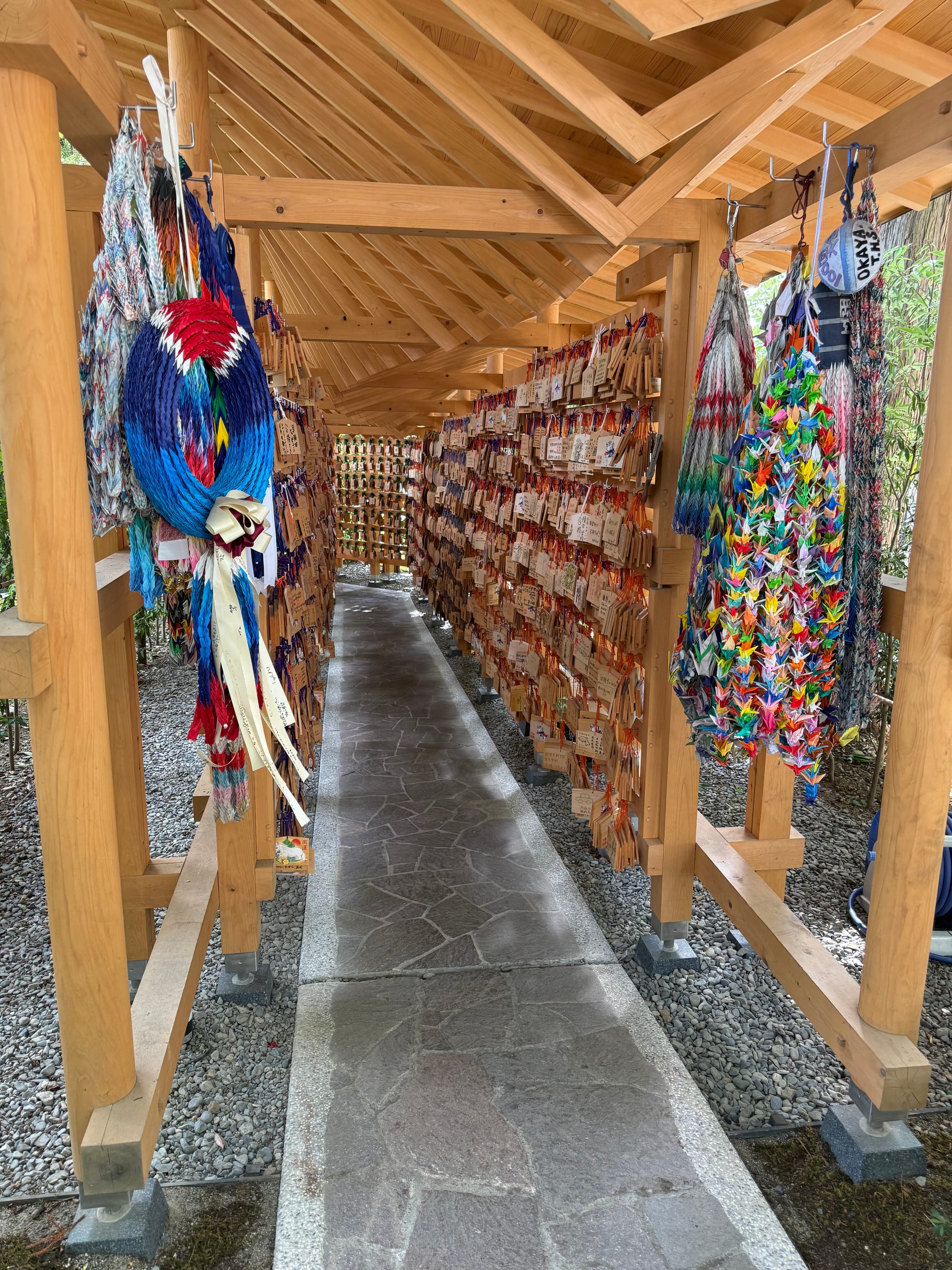 Covered walkway at Suwa Taisha Shimosha Akimiya lined with wooden prayer plaques and colourful folded-paper crane displays, creating a tunnel of wishes within the shrine grounds.