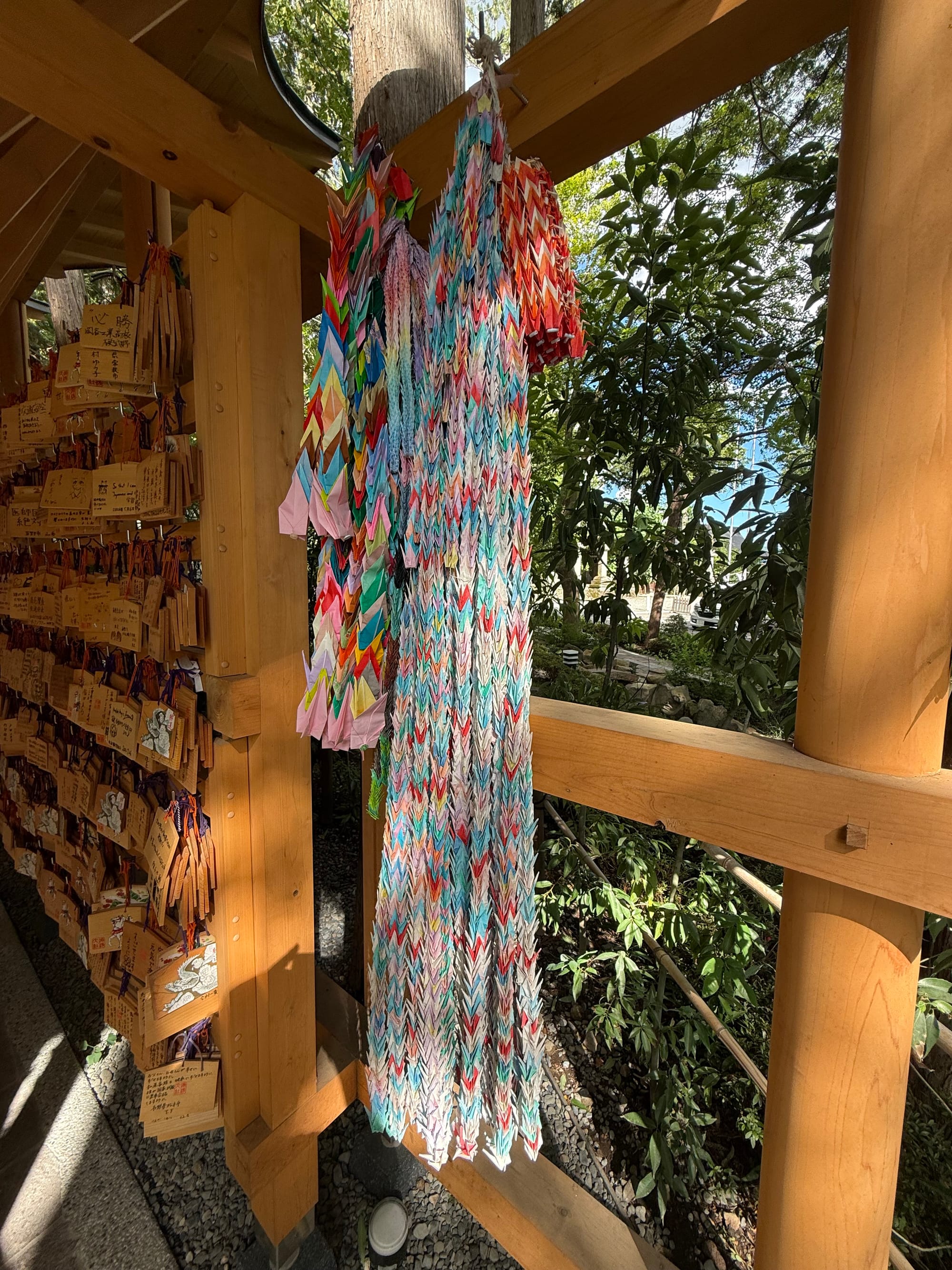 Colourful strings of folded paper cranes hang beside wooden prayer plaques at Suwa Taisha Shimosha Akimiya, displayed beneath a covered walkway within the shrine grounds.