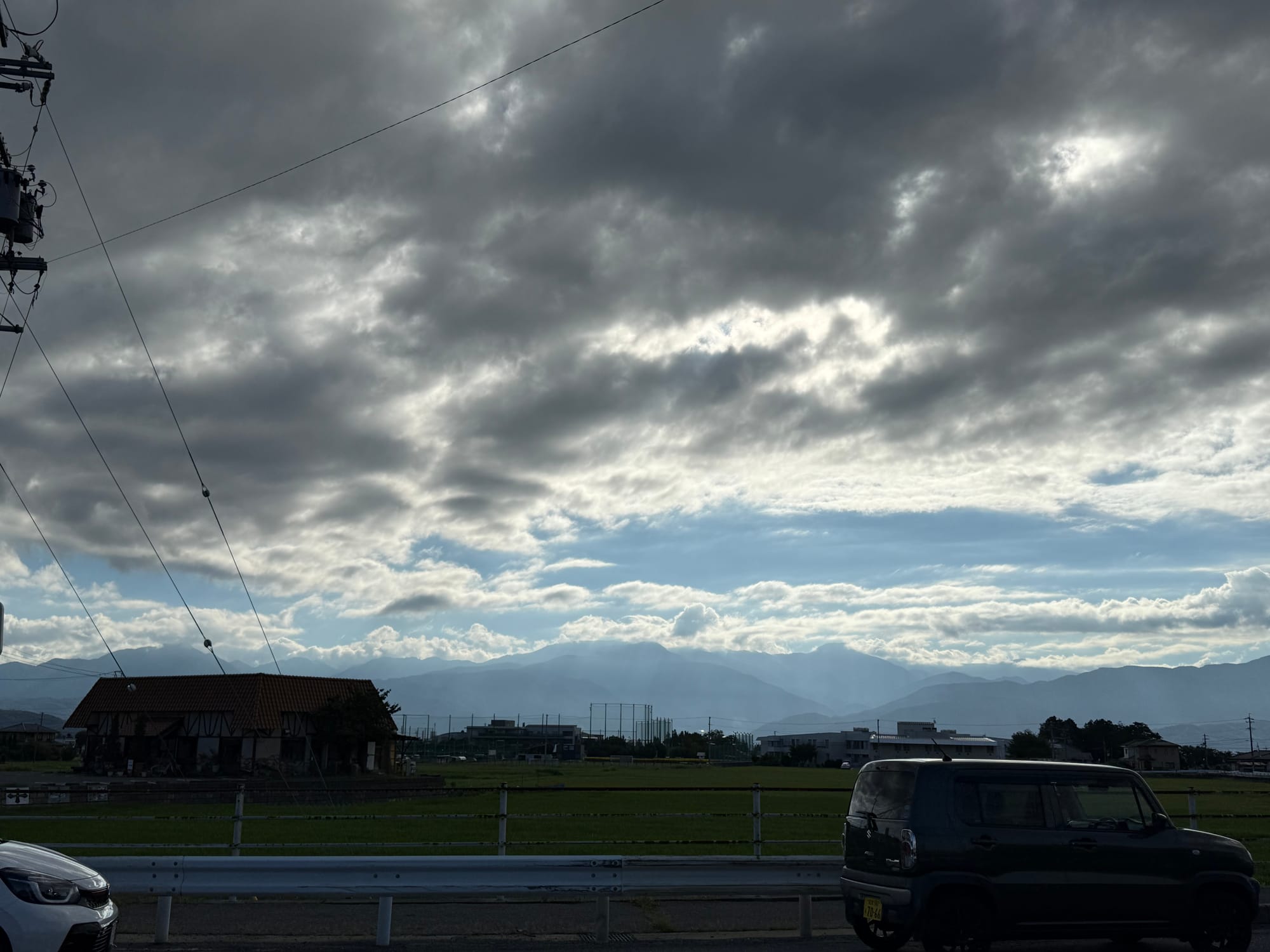 View from a car park near Matsumoto, showing fields, parked vehicles and distant mountain ranges beneath dramatic dark clouds with sunlight breaking through in the distance.