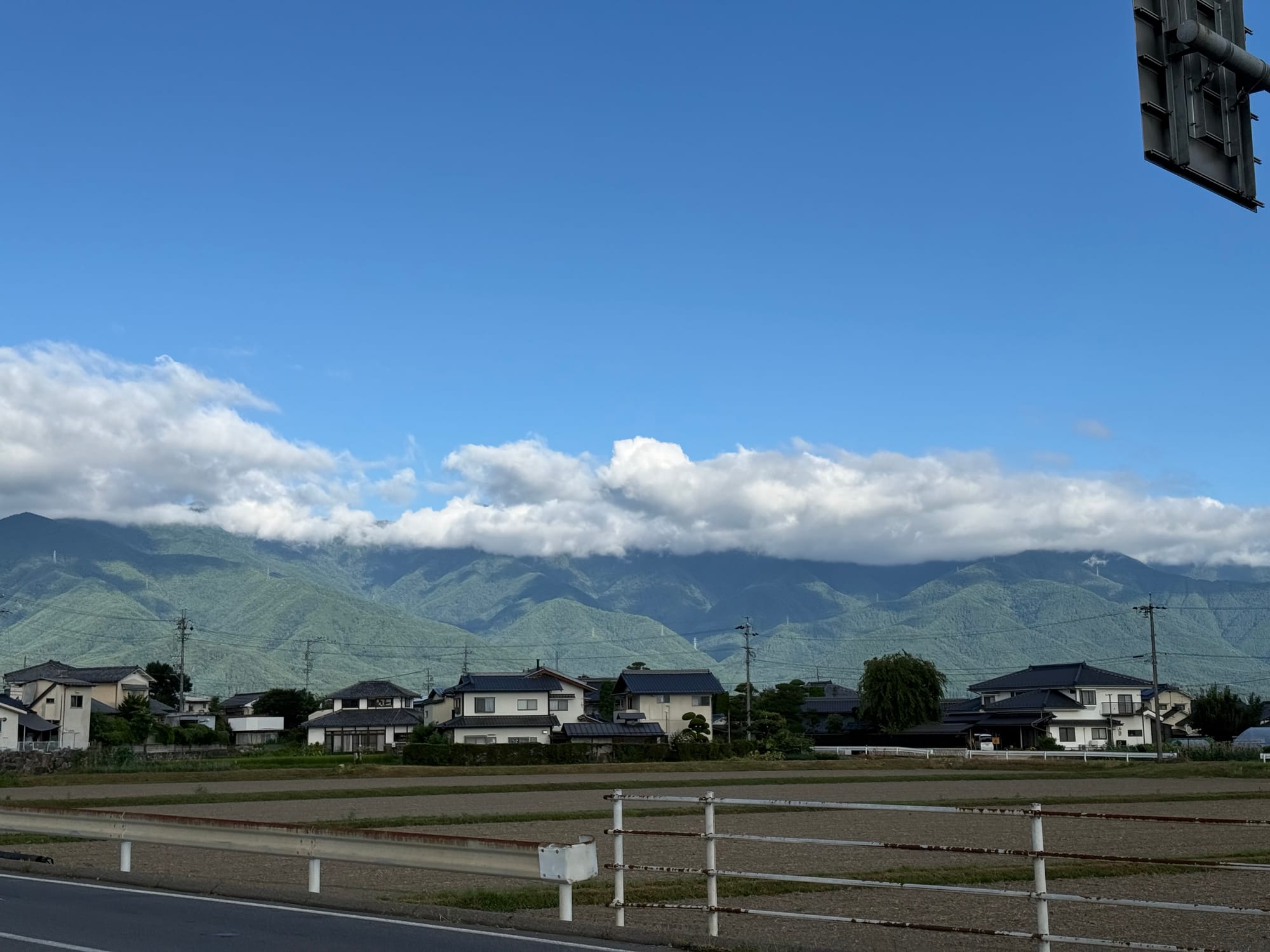 Mountain range near Matsumoto beneath a clear blue sky, with low clouds draped across the ridgeline above rural fields, houses and quiet roadside scenery.