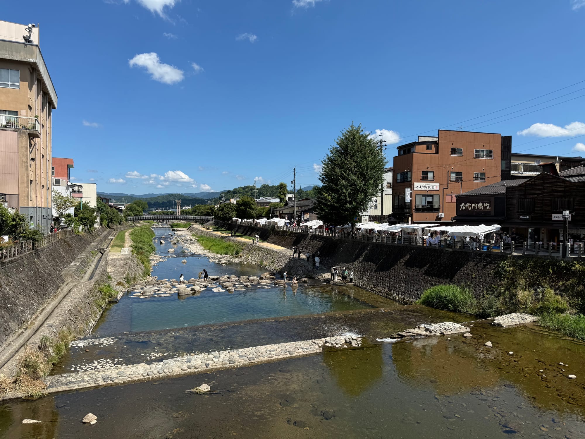 Miyagawa River flowing through central Takayama, with clear water, stone embankments, riverside cafés and traditional-style buildings beneath a bright blue sky.