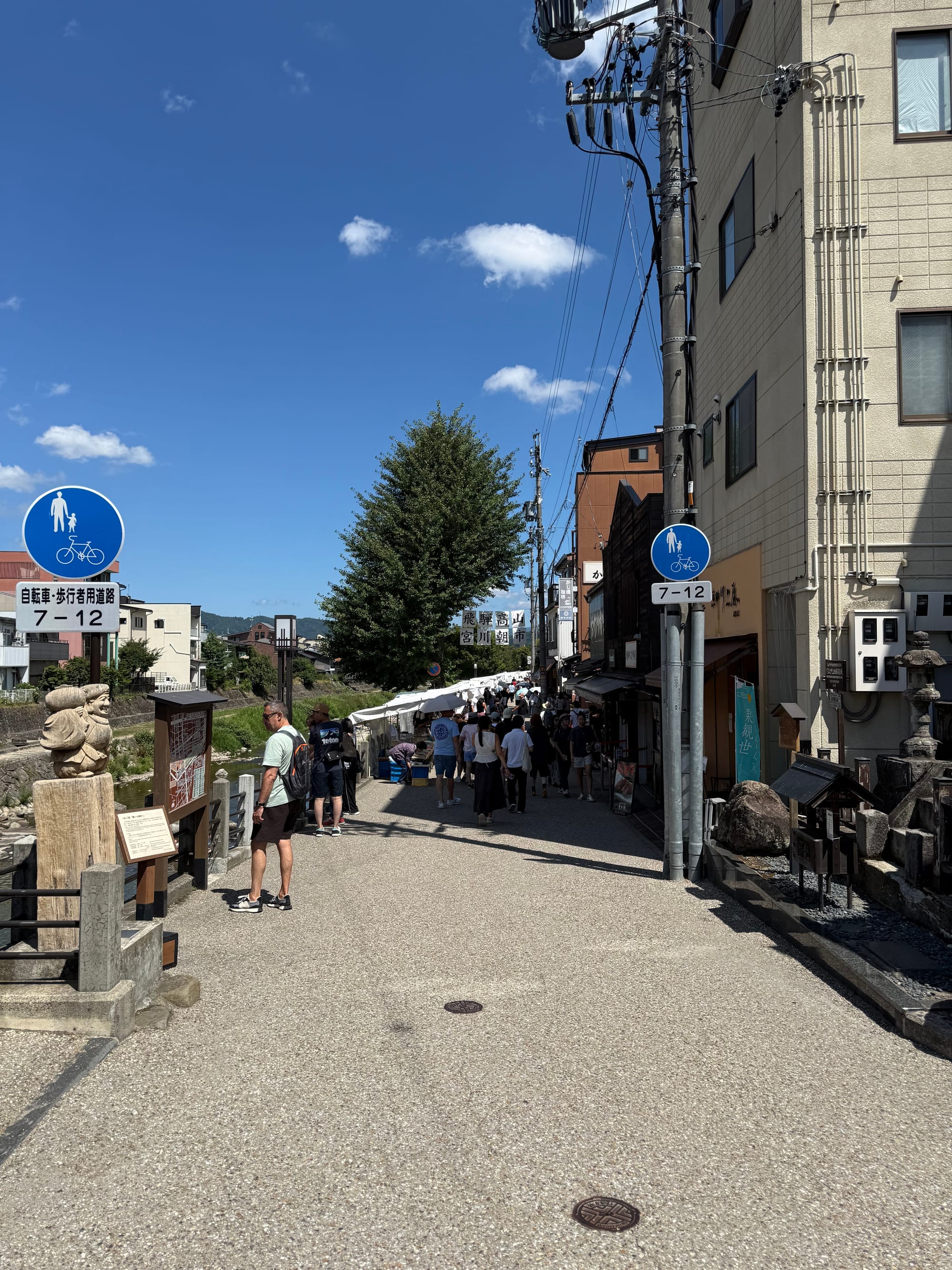Miyagawa Morning Market in Takayama, with visitors browsing stalls along the riverside walkway beneath white canopies on a sunny day in the historic city centre.