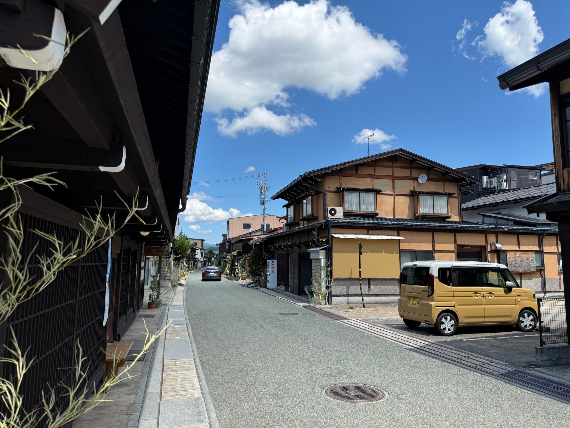 Traditional street in Takayama's historic district, lined with wooden merchant houses and parked vehicles beneath a bright blue sky with scattered summer clouds.