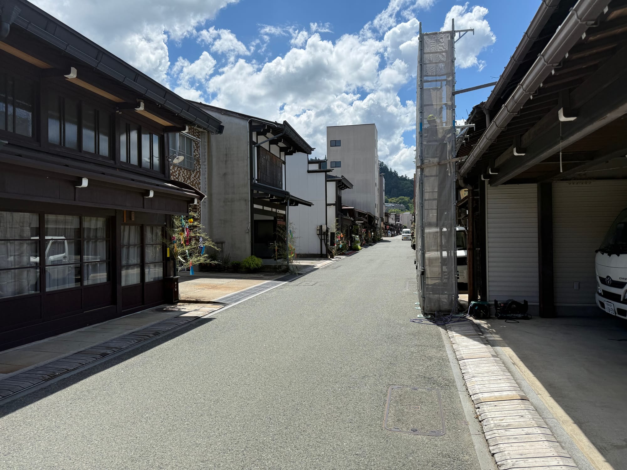 Street in Takayama's historic district lined with traditional wooden buildings, with summer decorations outside shopfronts and large white clouds drifting across a blue sky.