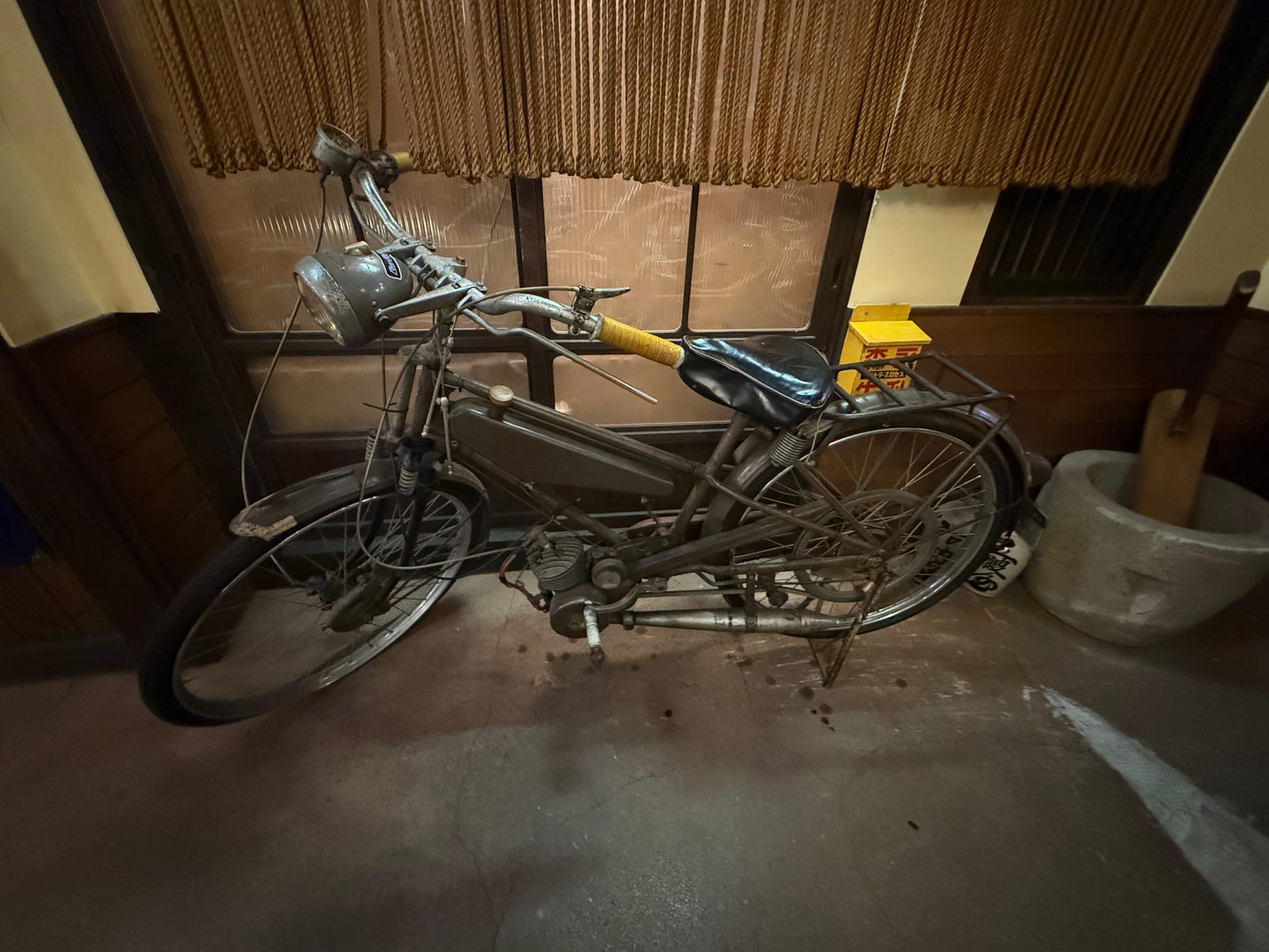 Vintage motorised bicycle displayed at the Takayama Showa-kan Museum, featuring a small engine mounted within the frame, a headlamp, rear rack and weathered components.