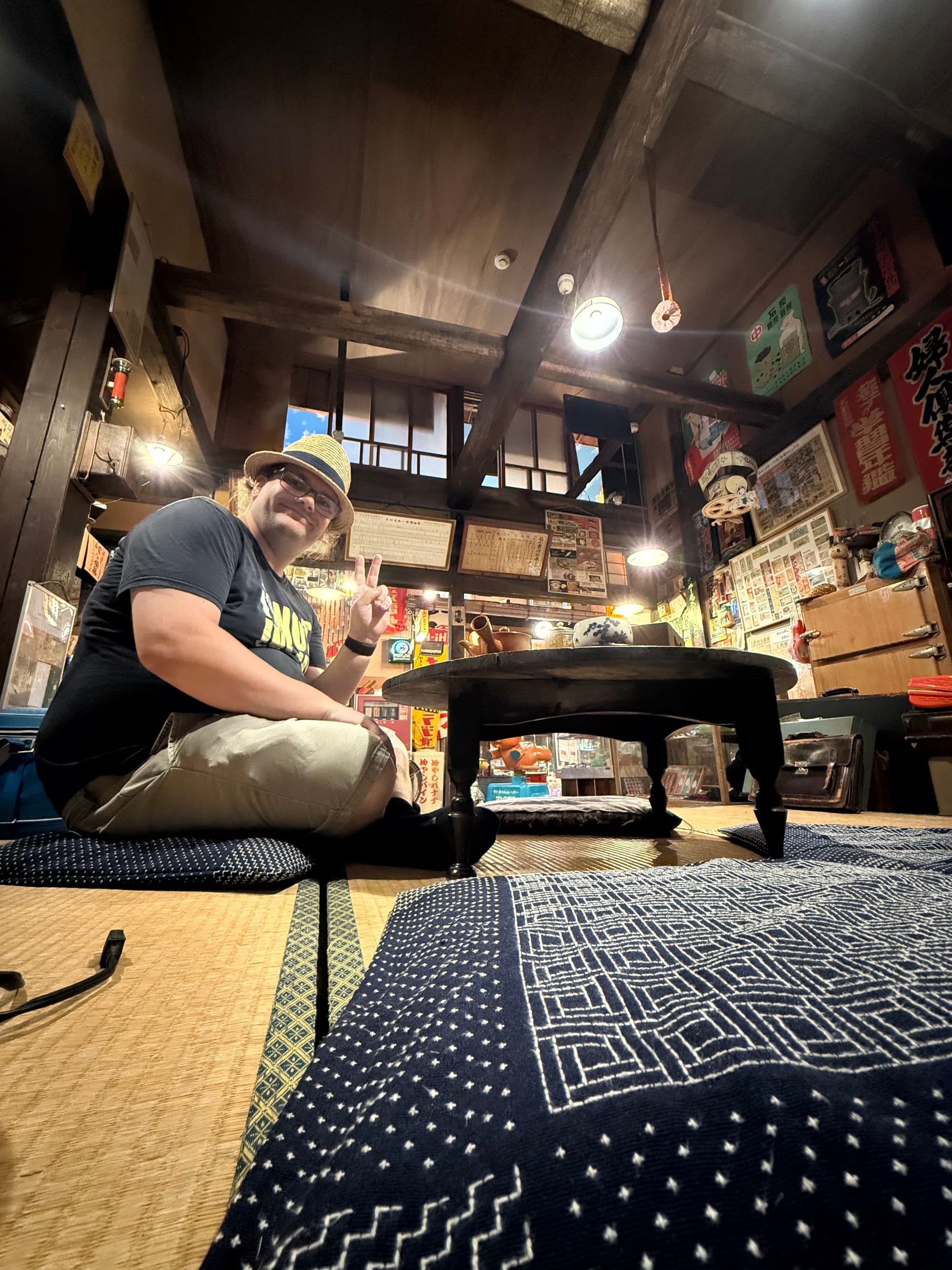 Visitor seated at a low table in a recreated Showa-era living room at the Takayama Showa-kan Museum, surrounded by vintage furnishings, advertisements and household items.