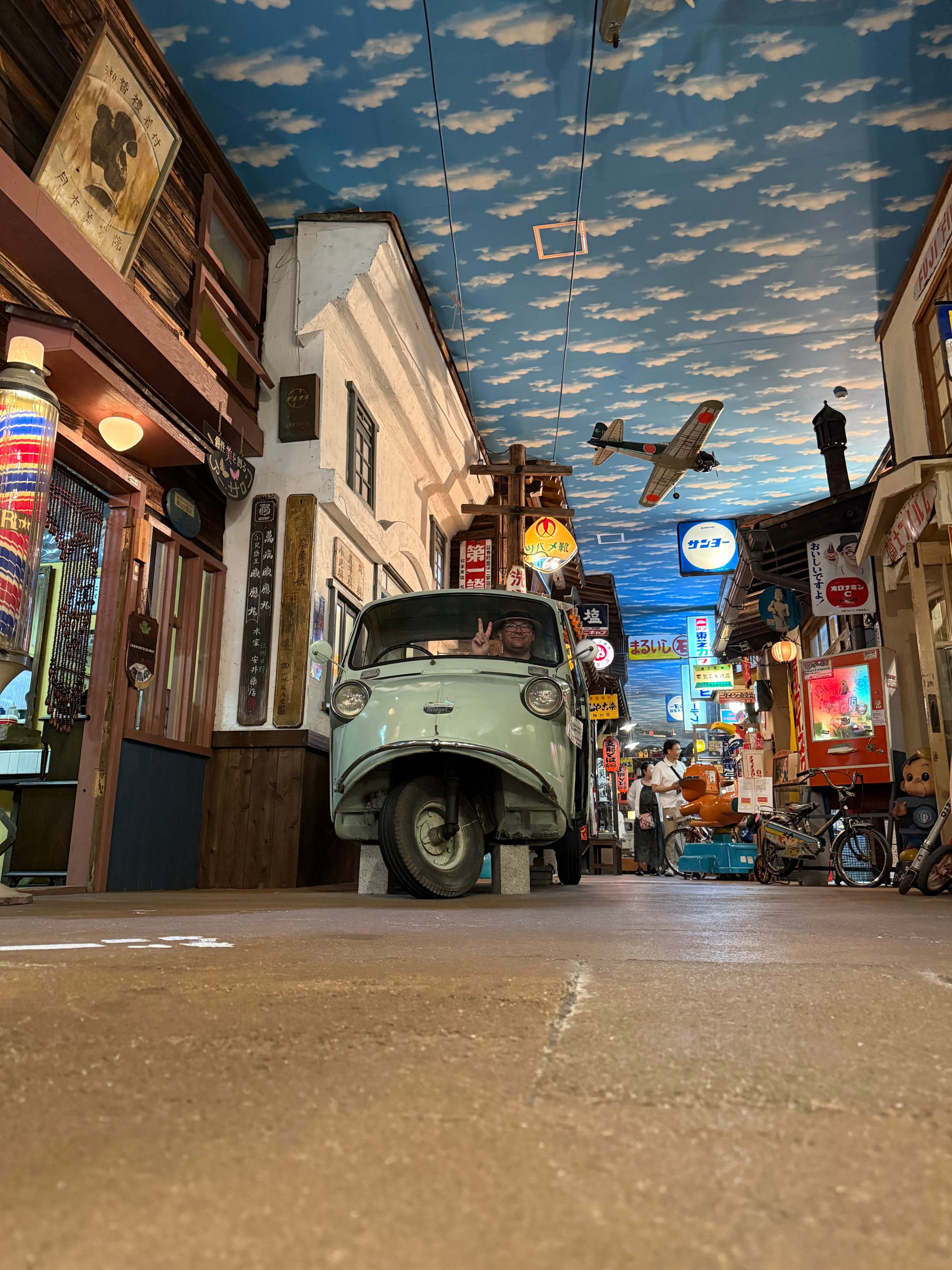 Daihatsu Midjet three-wheeled utility vehicle displayed on a recreated Showa-era street at the Takayama Showa-kan Museum, with retro shopfronts, signs and a model aircraft overhead.