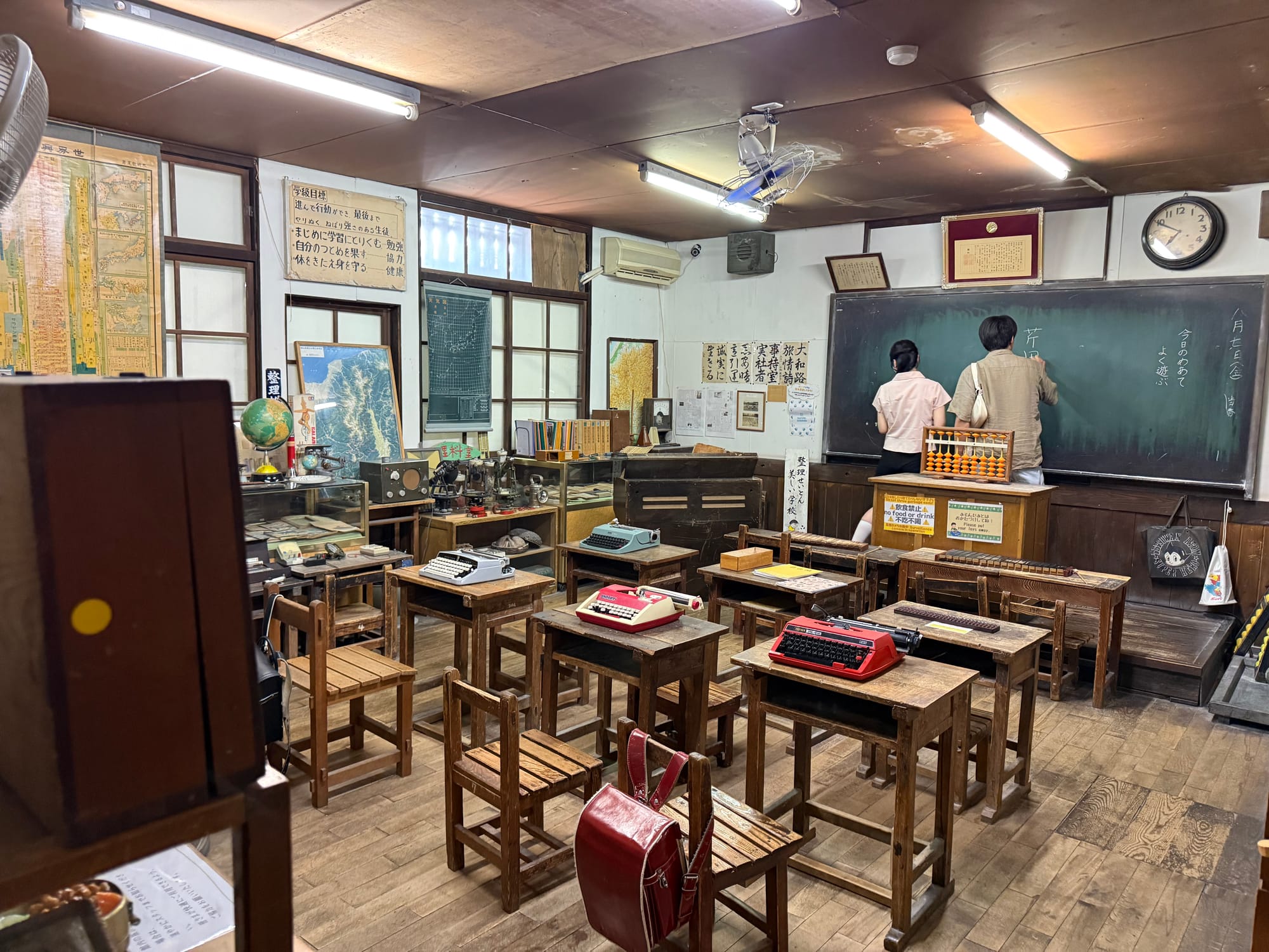 Recreated Showa-era classroom at the Takayama Showa-kan Museum, featuring wooden desks, typewriters, maps, an abacus, a blackboard and period educational materials.