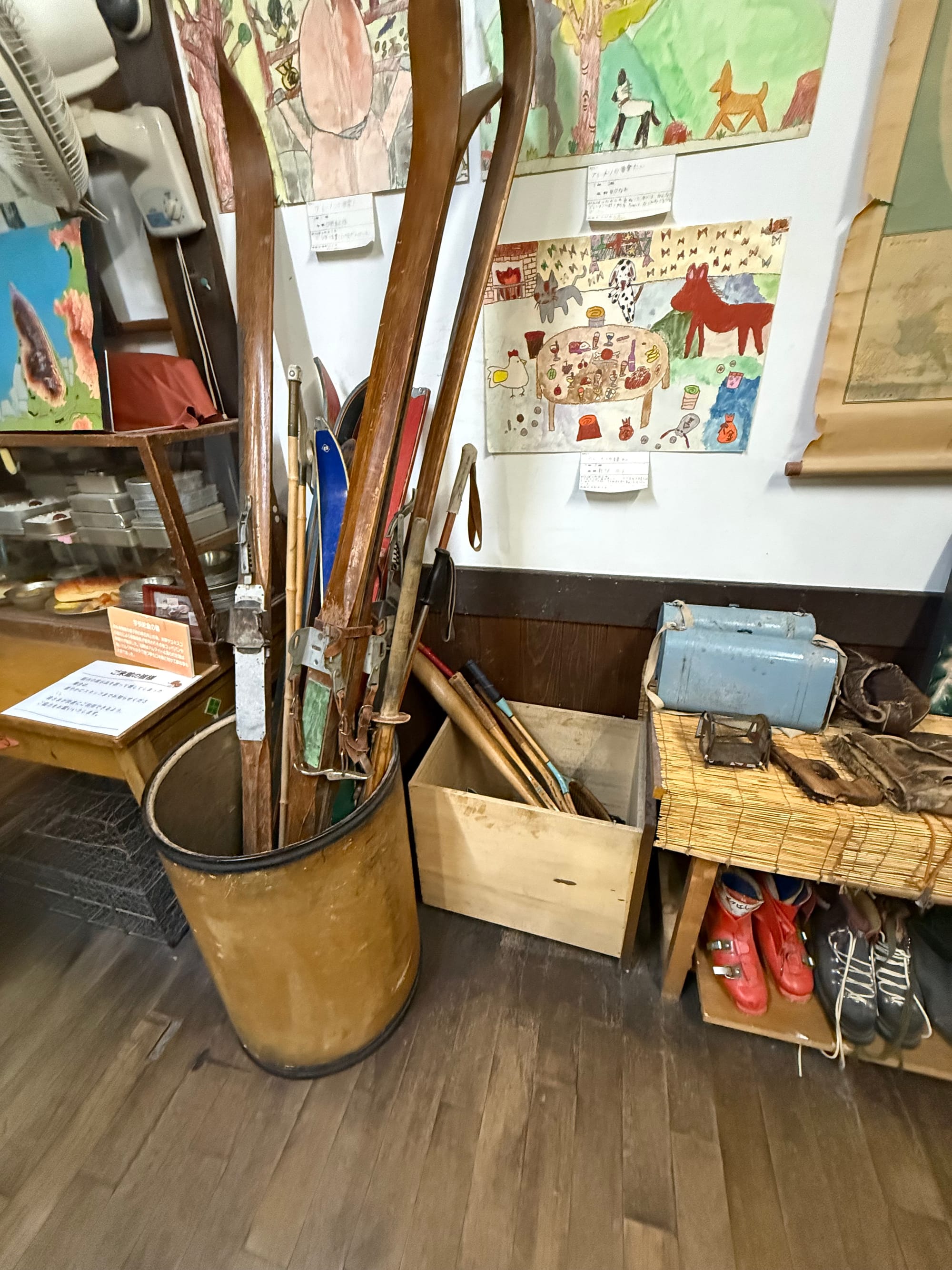 Vintage skis, ski boots and winter sports equipment displayed in a recreated Showa-era classroom at the Takayama Showa-kan Museum, alongside children's artwork and school items.