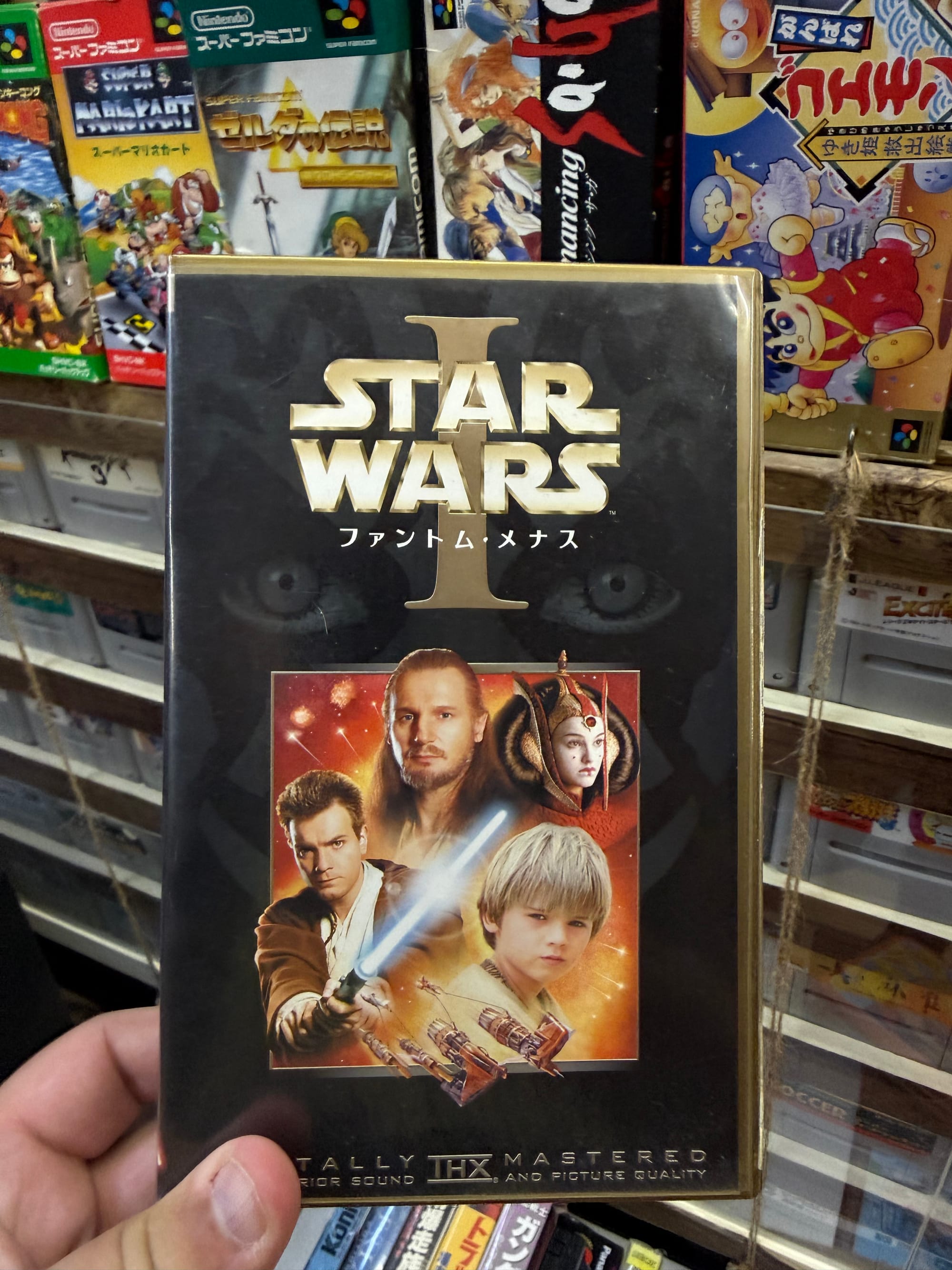 Japanese VHS release of Star Wars Episode I: The Phantom Menace displayed at the Takayama Showa-kan Museum, held in front of shelves filled with retro video games and media.