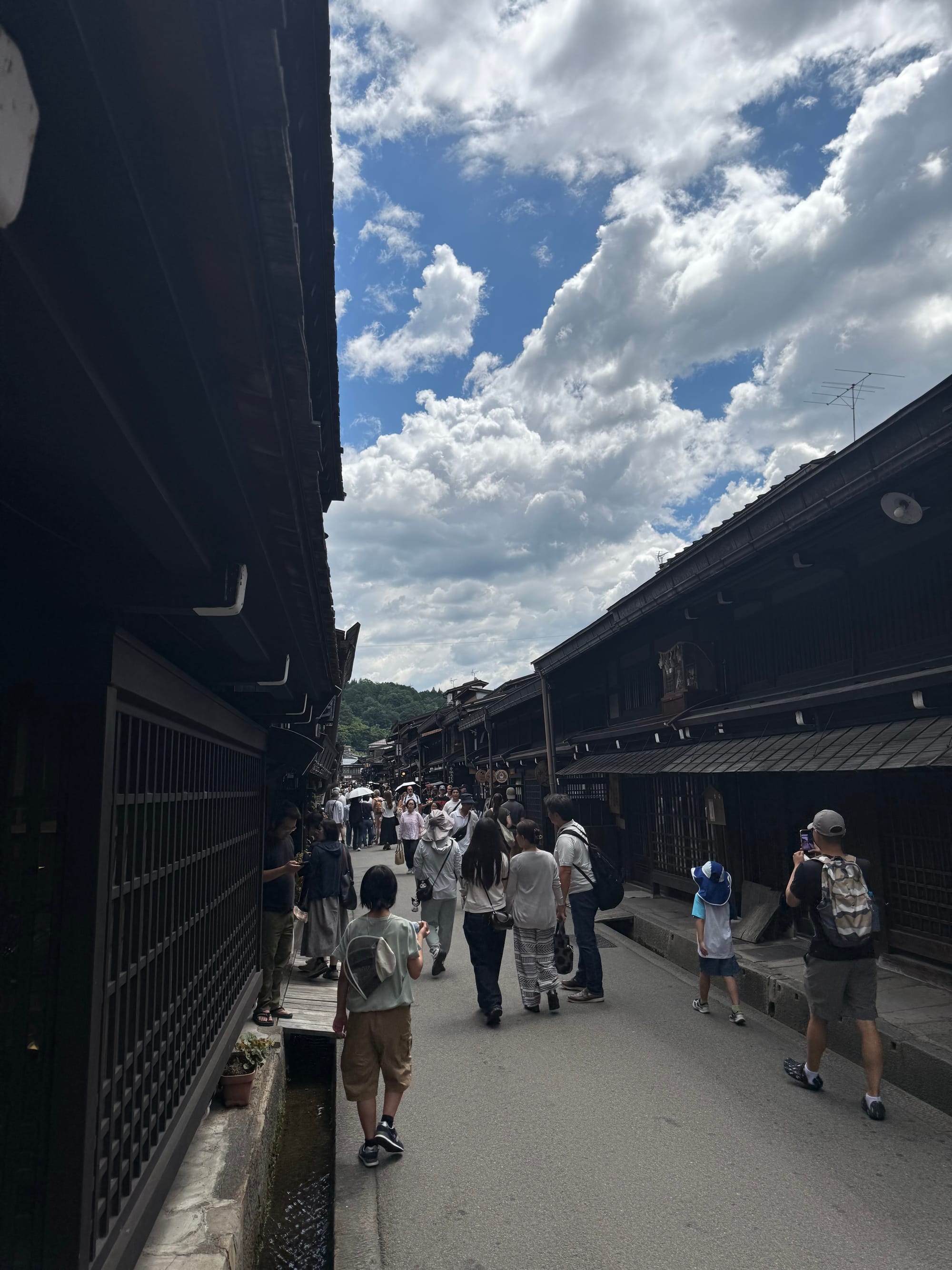 Visitors walk along a street in Takayama's historic old town, lined with traditional wooden merchant houses beneath a dramatic sky filled with towering summer clouds.