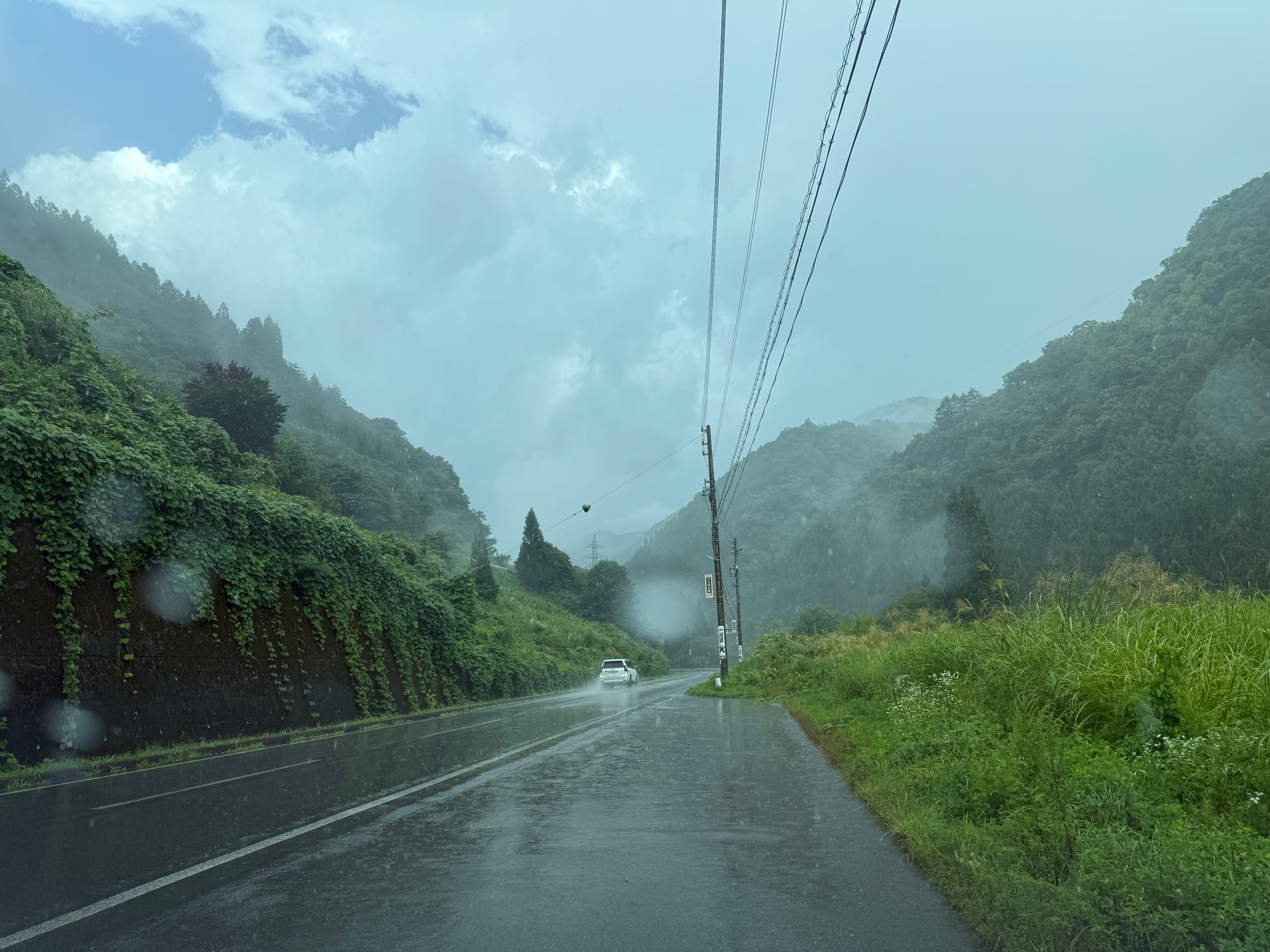 Rain falls on a mountain road in central Japan, with wet pavement, mist-covered forested hills, power lines and a vehicle driving through a heavy summer downpour.