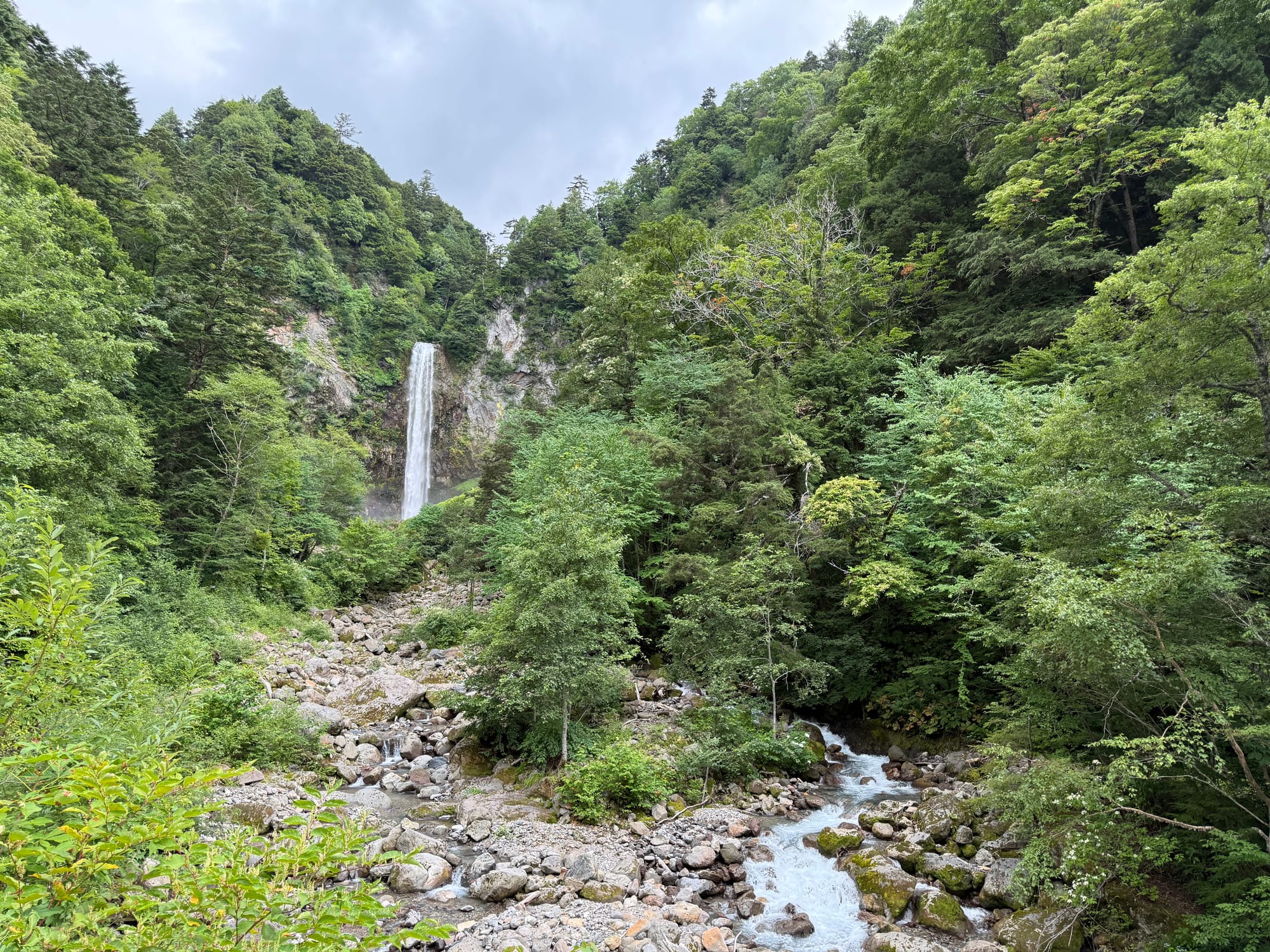Hirayu Great Falls cascading down a rocky cliff in the Northern Japan Alps, surrounded by dense forest, with a mountain stream flowing through the boulder-strewn valley below.
