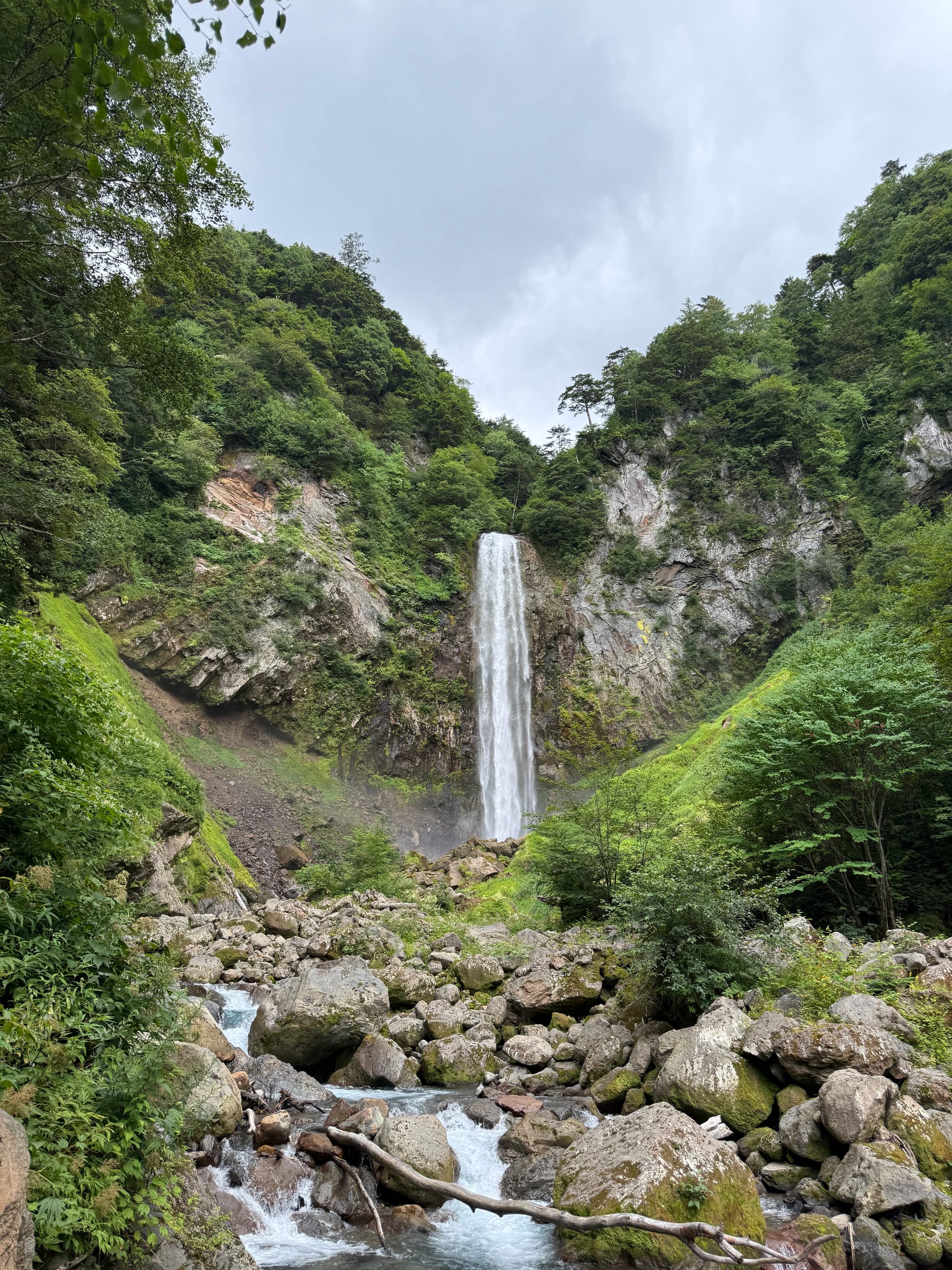 Hirayu Great Falls plunging into a rocky basin surrounded by lush forest, with a mountain stream flowing through large boulders at the base of the waterfall.
