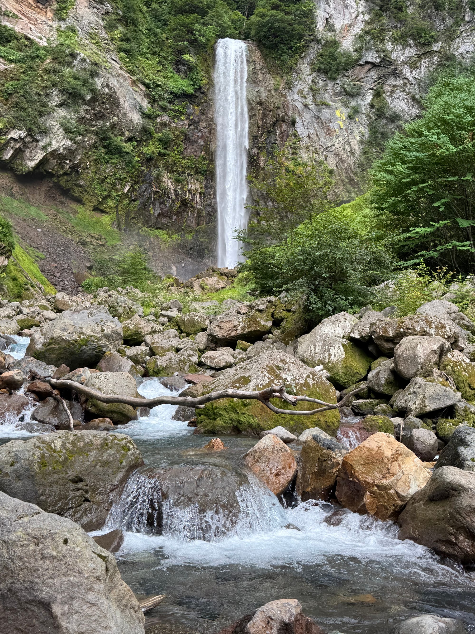 Hirayu Great Falls viewed from its rocky base, with water plunging down a steep cliff into a mountain stream flowing through large boulders in a forested alpine valley.