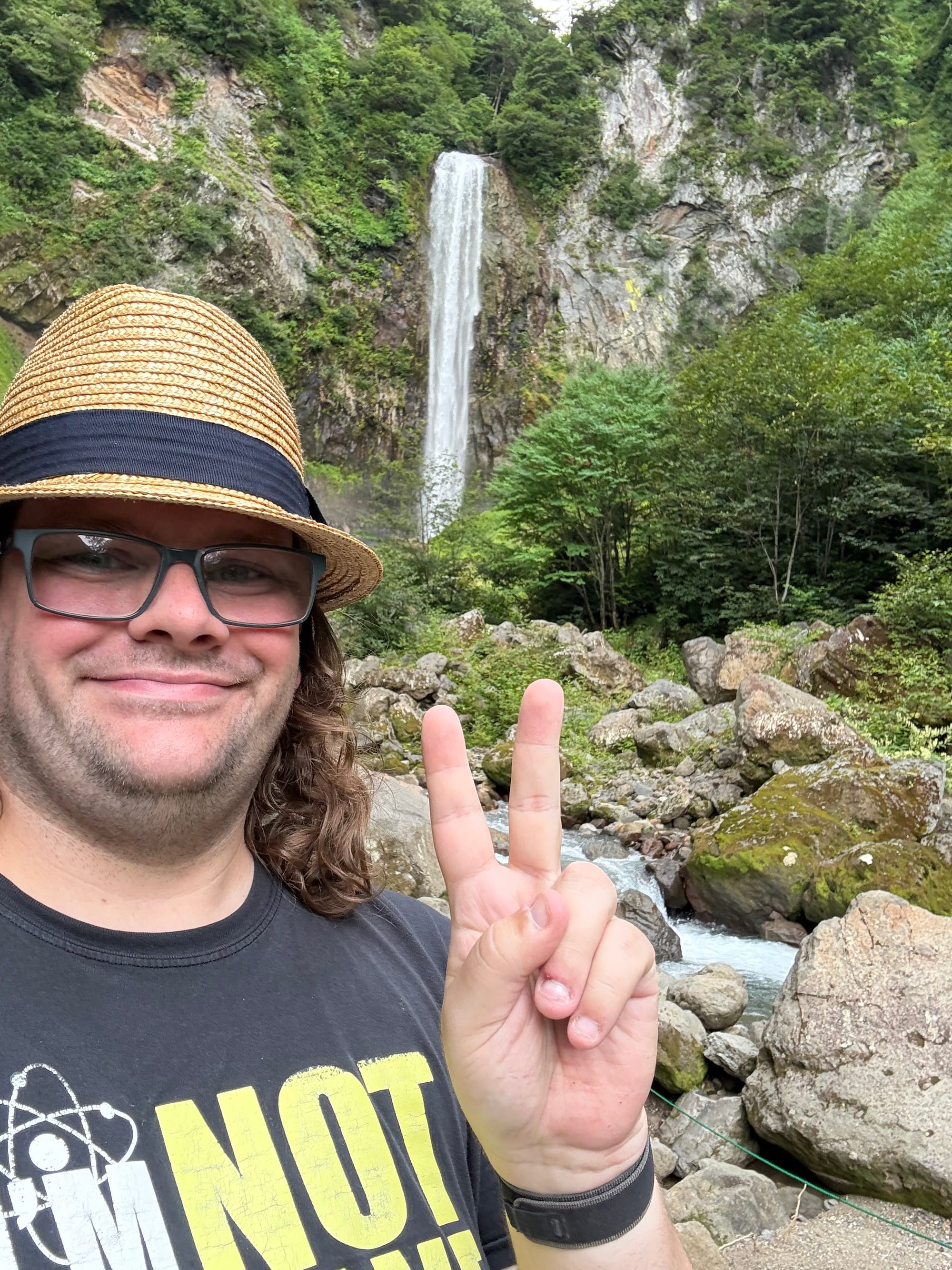 Visitor posing beside Hirayu Great Falls, making a peace sign in front of the waterfall, with a rocky stream, forested cliffs and alpine scenery in the background.