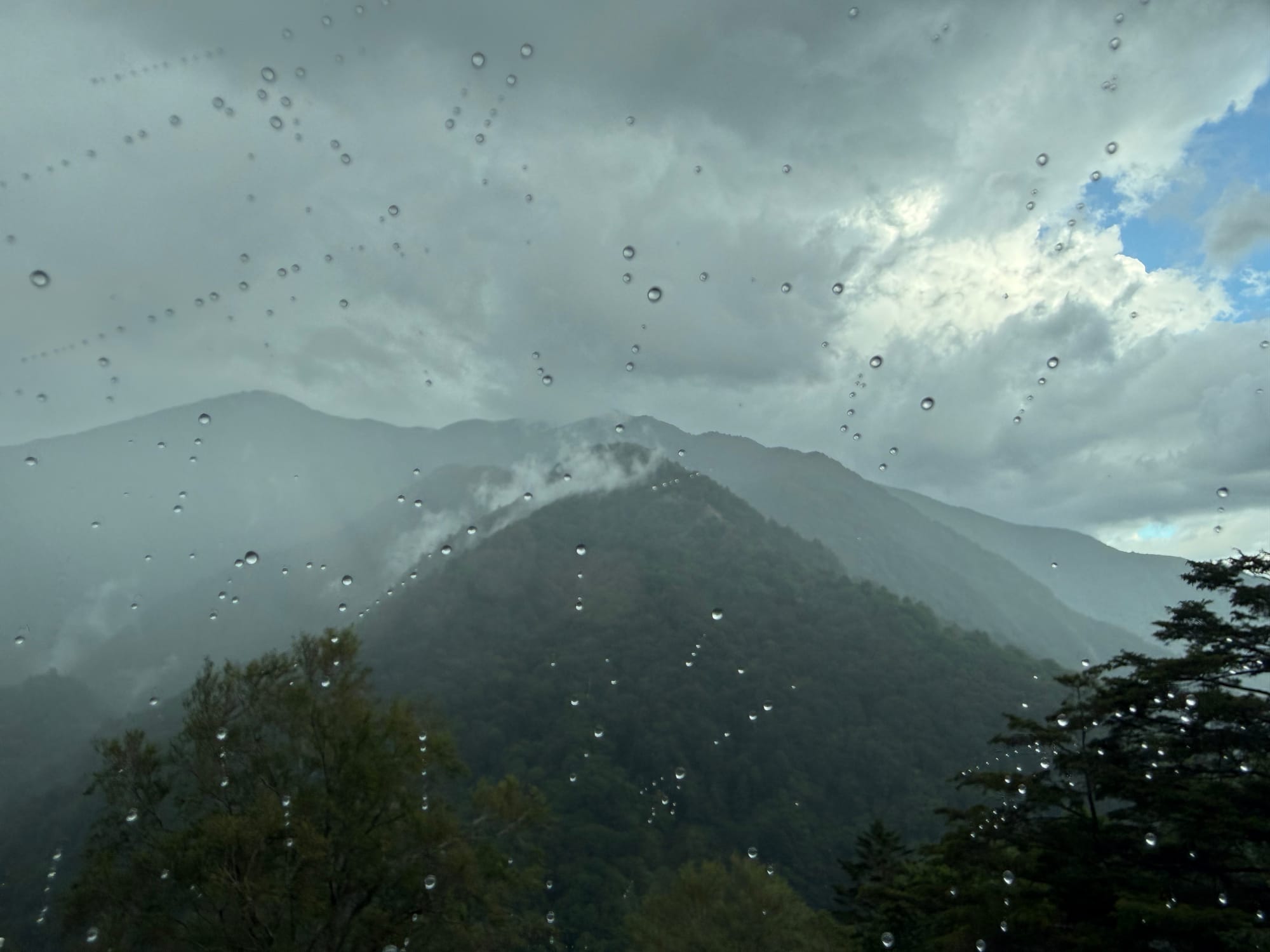 Rain-soaked mountain landscape viewed through a car window at Abo Pass, with raindrops on the glass, mist rising from forested slopes and storm clouds over the Alps.
