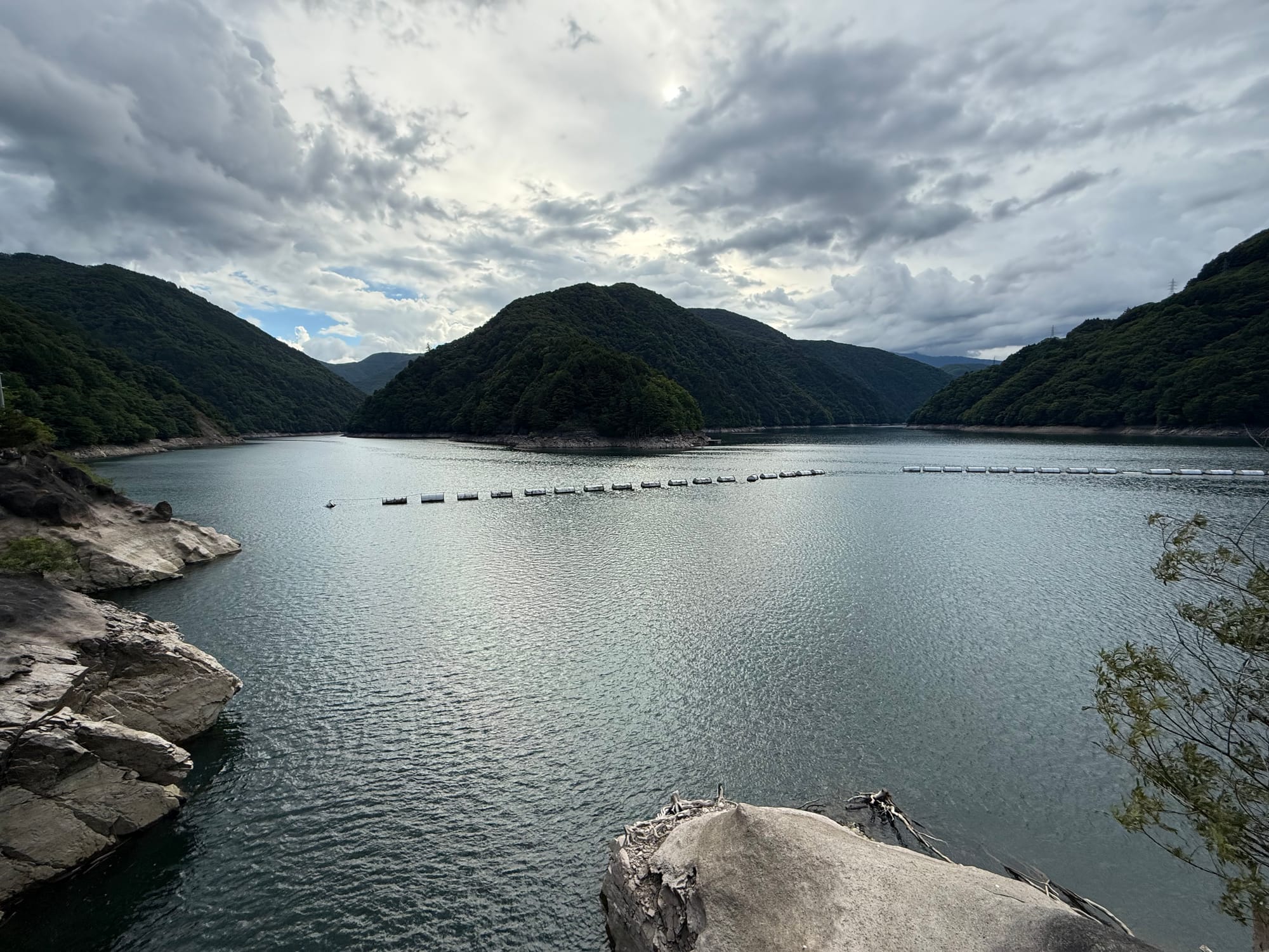 Nagawado Dam reservoir surrounded by forested mountains, with floating debris barriers crossing the water and dramatic storm clouds gathering above the Azusa River valley.