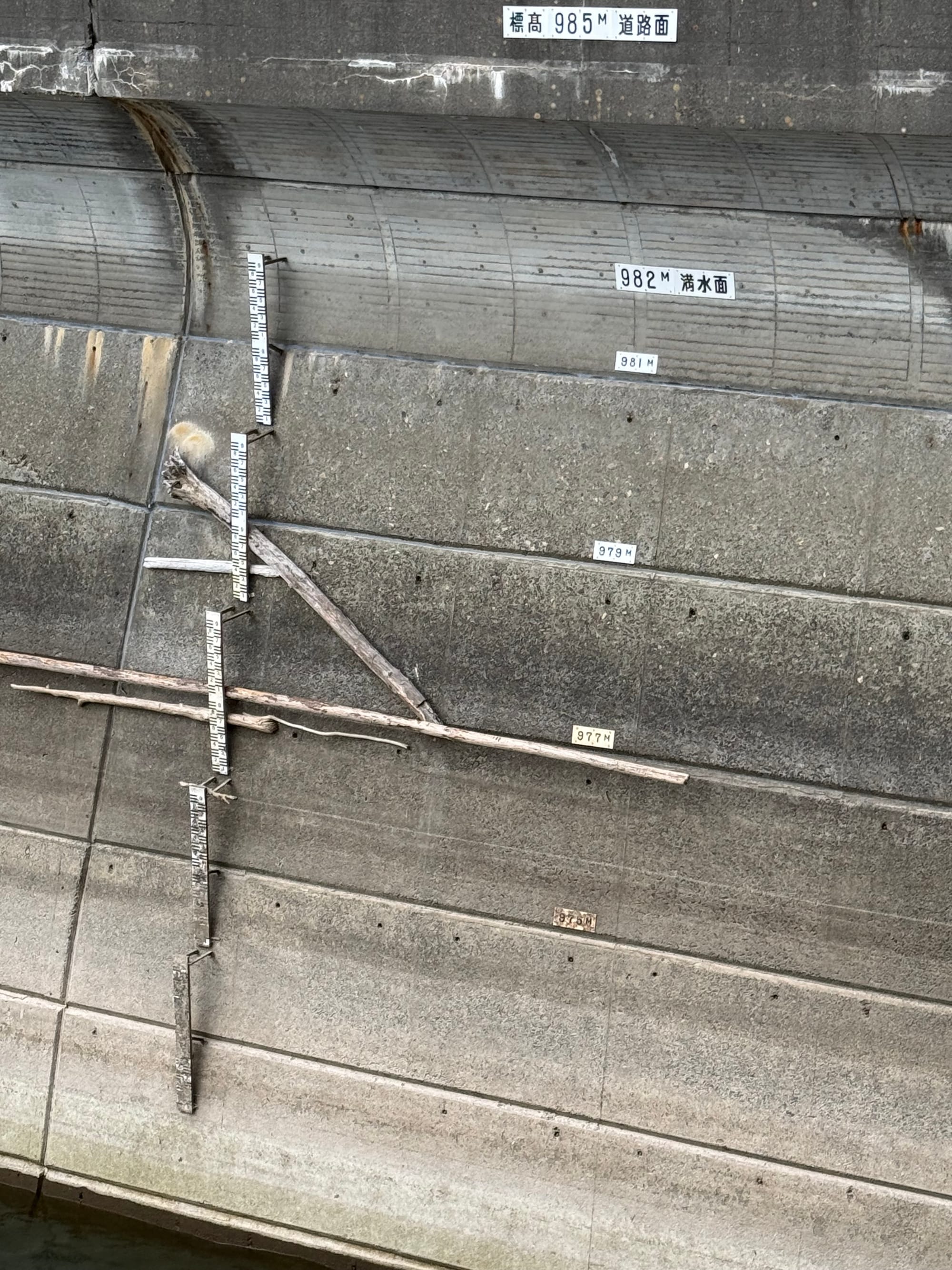 Water level gauge and elevation markings on the concrete face of Nagawado Dam, showing reservoir levels in metres above sea level with driftwood lodged against the structure.